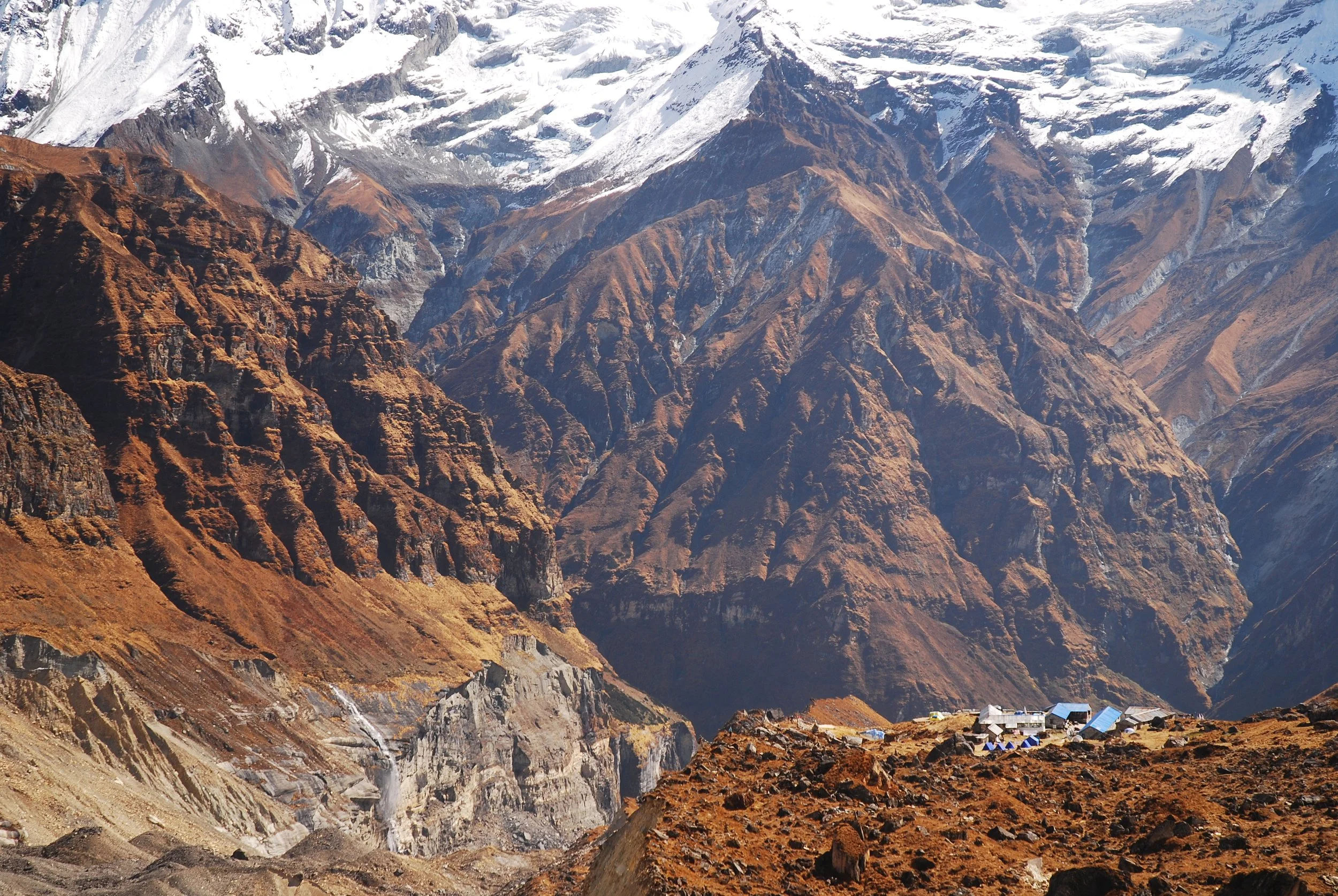 A mountain landscape with steep brown and gray rocky slopes, snow-capped peaks in the distance, and a small village with buildings and blue roofs at the base of the mountains.