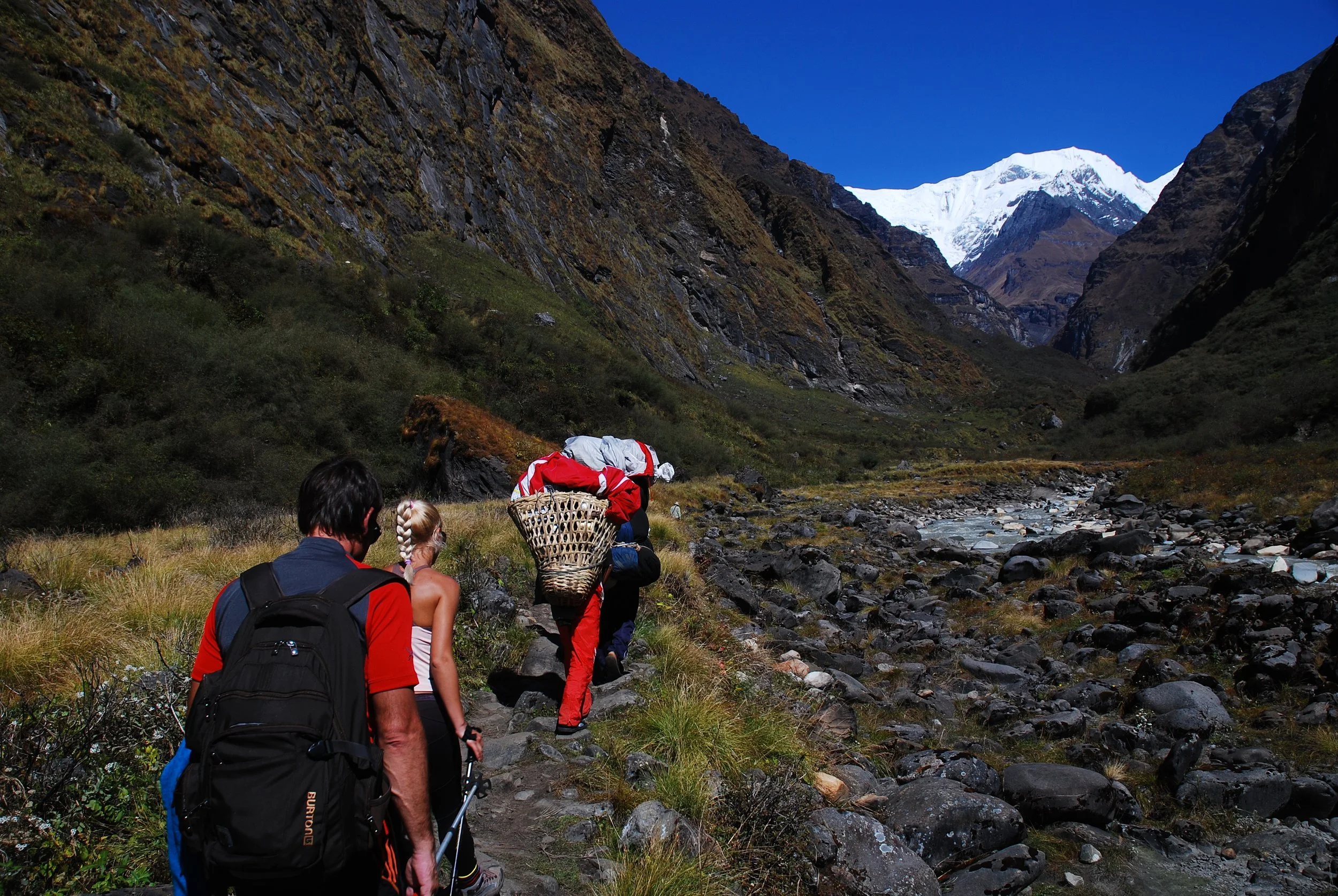 A group of hikers trekking through a mountainous landscape with snow-capped peaks in the background, rocky terrain, and a small stream on the side.