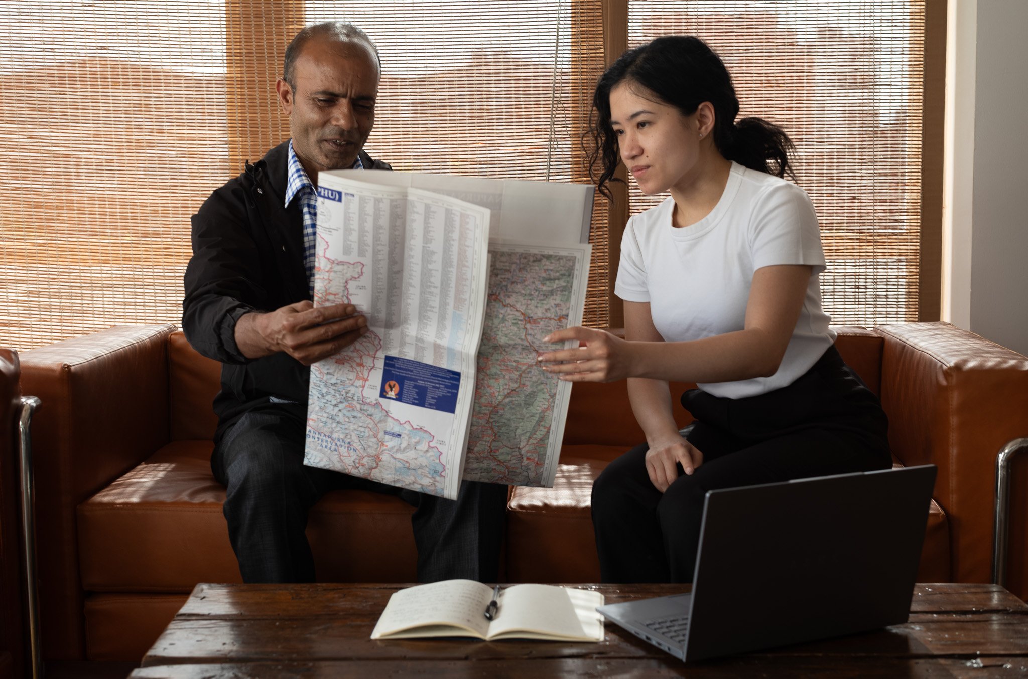A man and woman sitting on a couch in front of a window with blinds, looking at a large map. The man is holding the map, and the woman is reaching towards it. A laptop, an open notebook, and a pen are on the wooden table in front of them.
