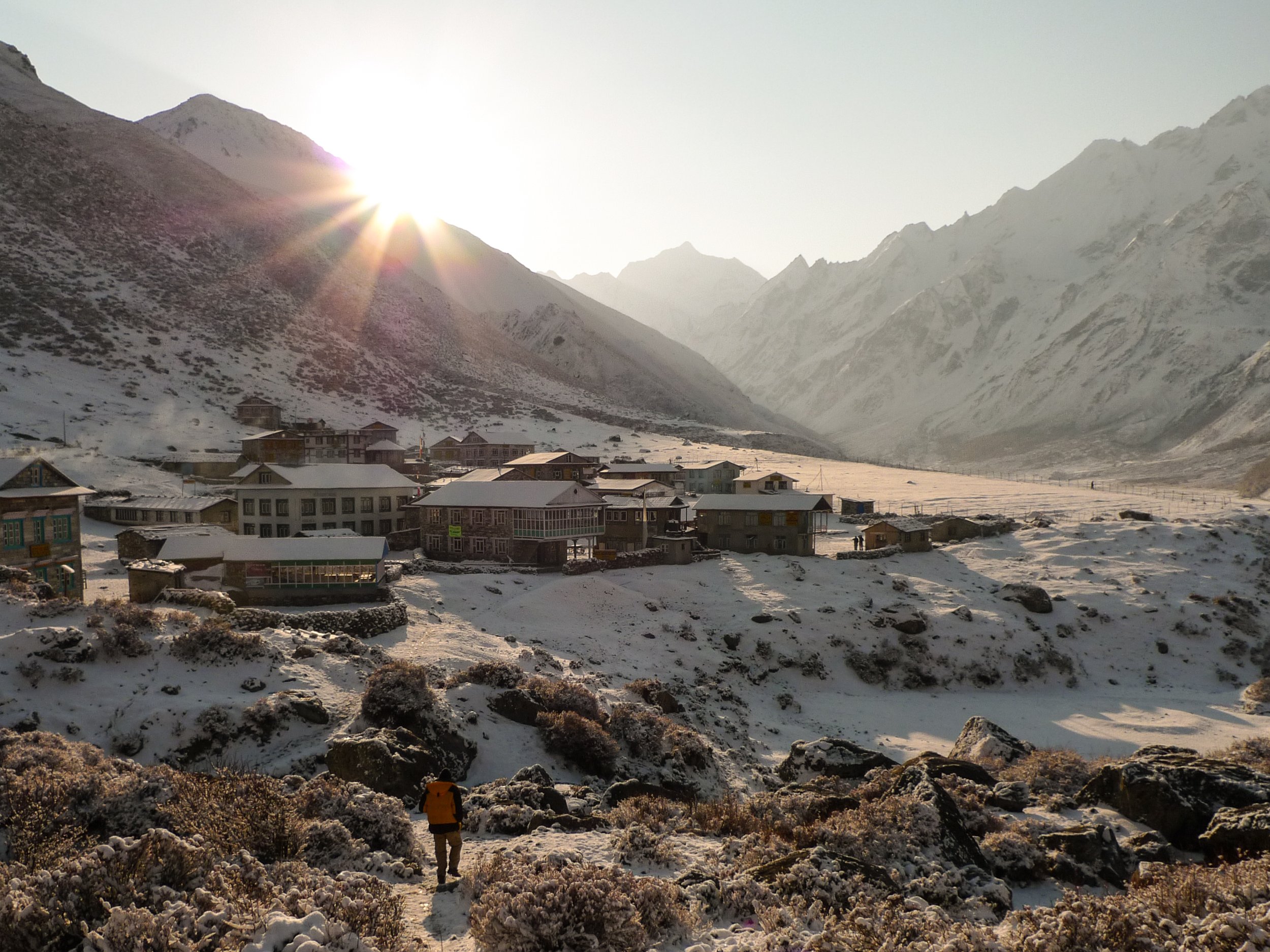 Snow-covered village in a mountain valley with mountains in the background, a person walking on a snowy path, and the sun rising behind the mountain.