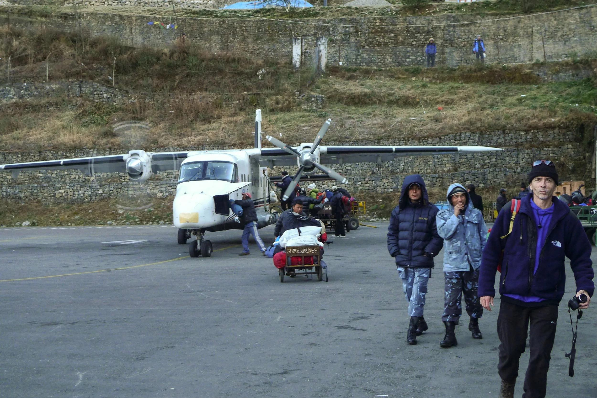 People boarding or disembarking a small propeller airplane on a tarmac in Lukla in Nepal, with some individuals walking and others handling luggage, set against a hillside background.