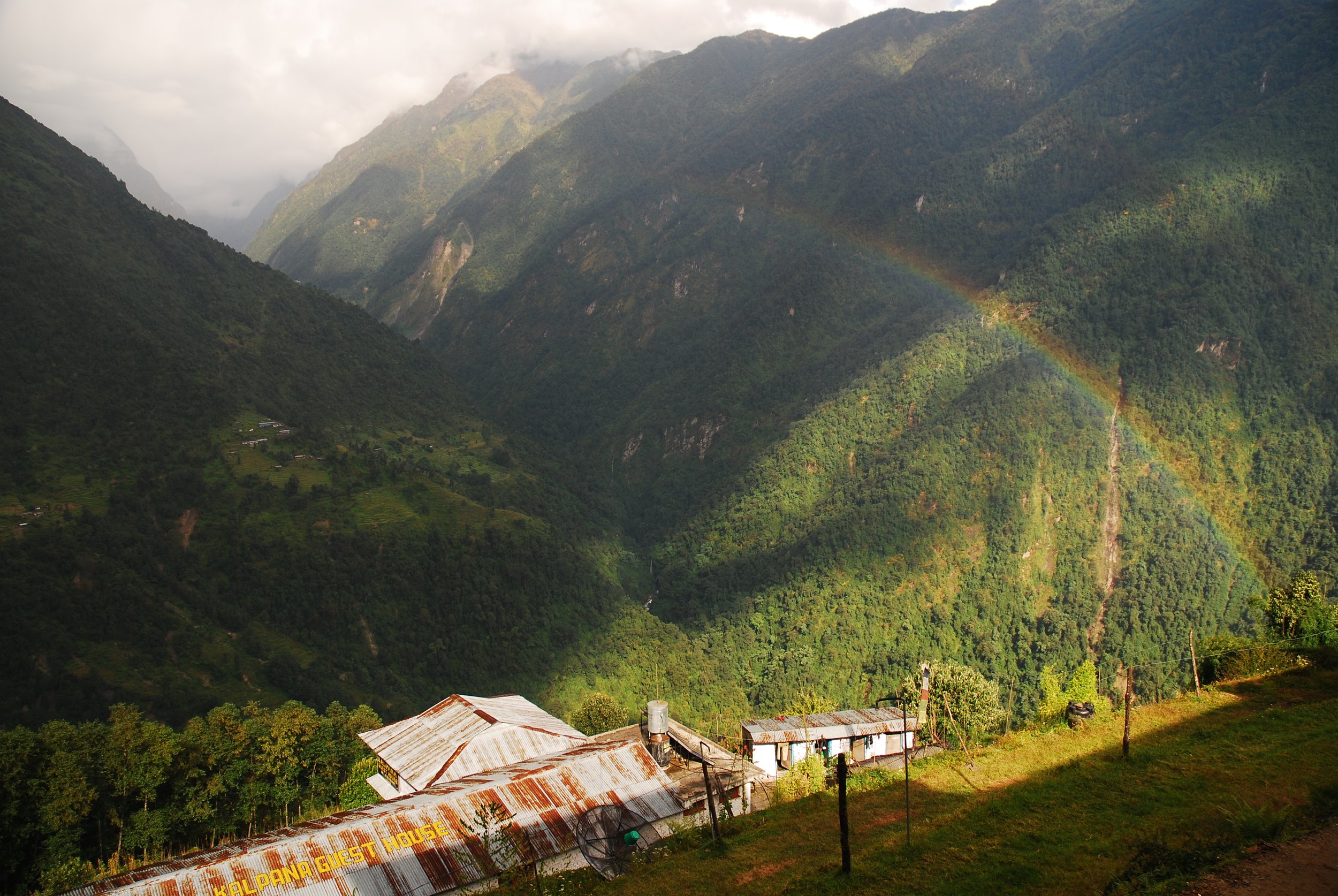 Rural village with rusted metal roofs and lush green hills, featuring a rainbow across the mountains in the background.
