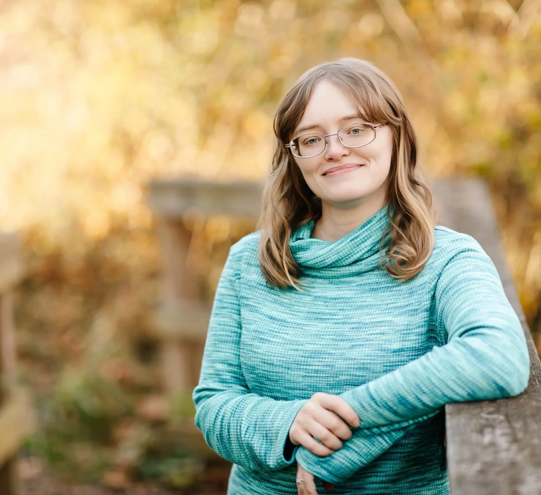 An adult woman with blond hair and glasses wearing a blue, long sleeved shirt. She is leaning against a wooden railing with autumn trees in the background.