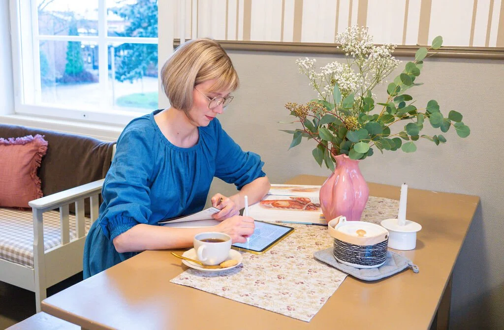 Syöpälääkäri pöydän ääressä selaamassa tablettia, vieressä teekuppi ja eukalyptuksen oksia maljakossa