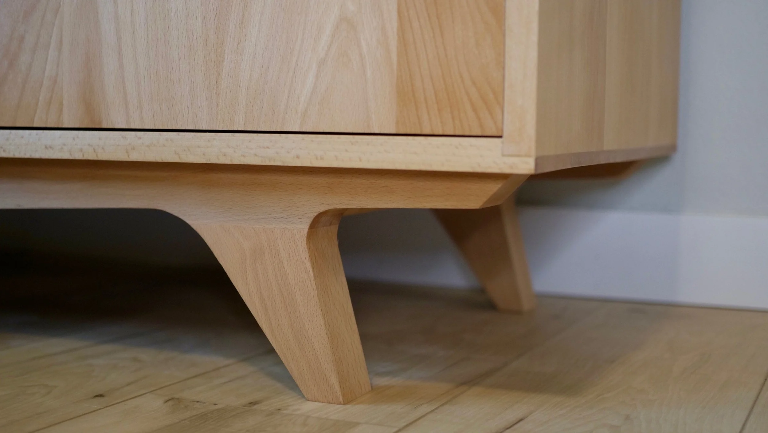Close-up of wooden furniture leg and part of the cabinet or drawer, with a wooden floor and light-colored wall in the background.