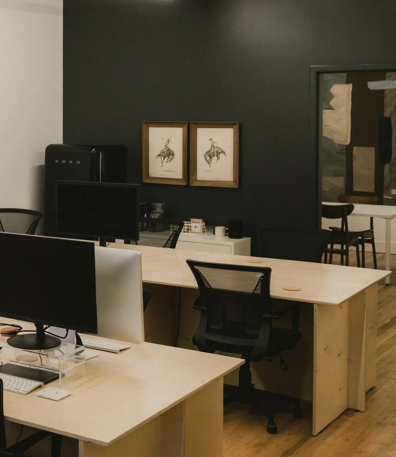 Modern office space with light wood desks, black ergonomic chairs, a flat-screen monitor, and dark accent wall with two framed black-and-white art pieces of horse riders.