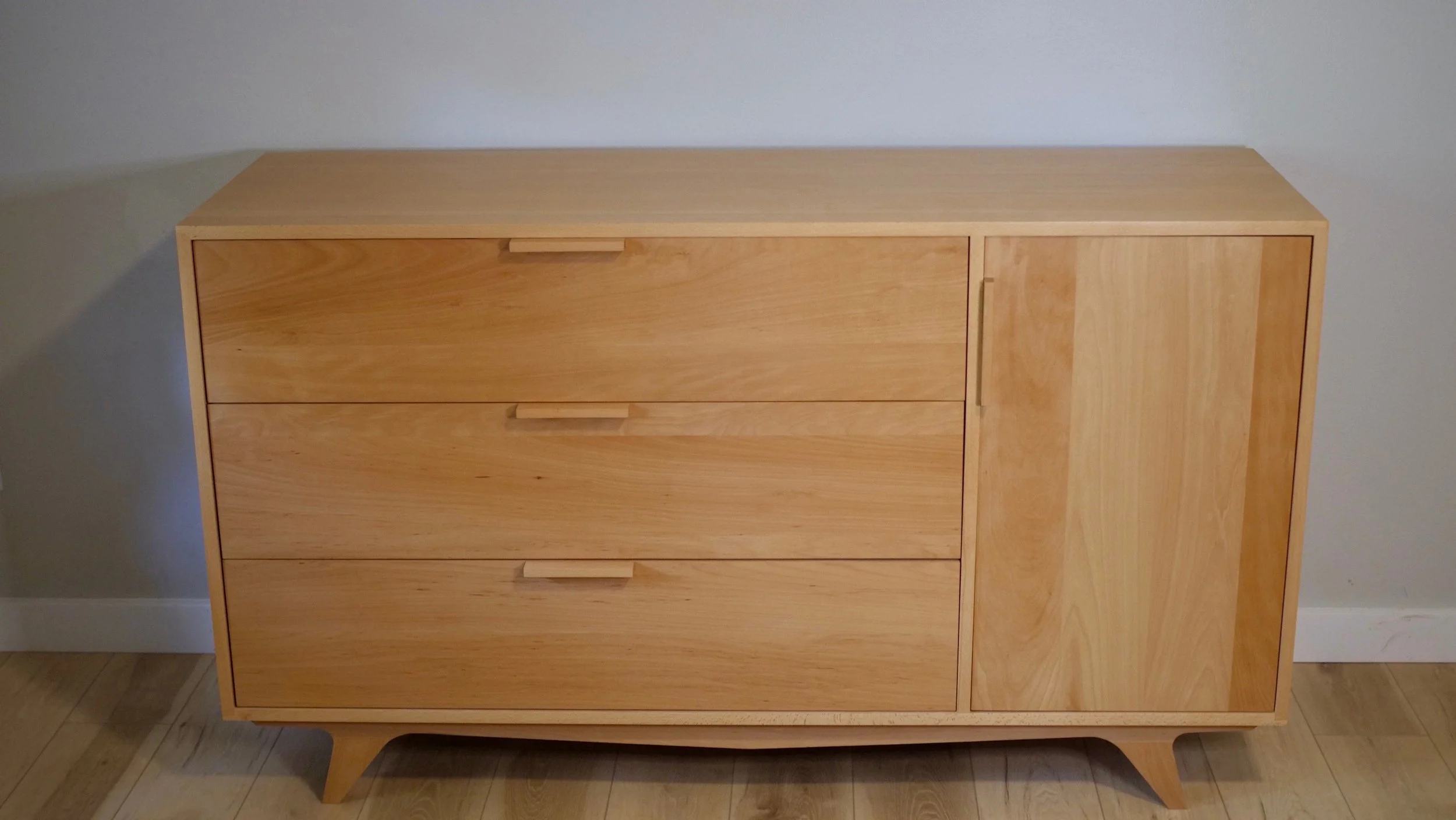 A light wood dresser with four drawers and a cabinet, standing on four angled legs, set against a white wall.