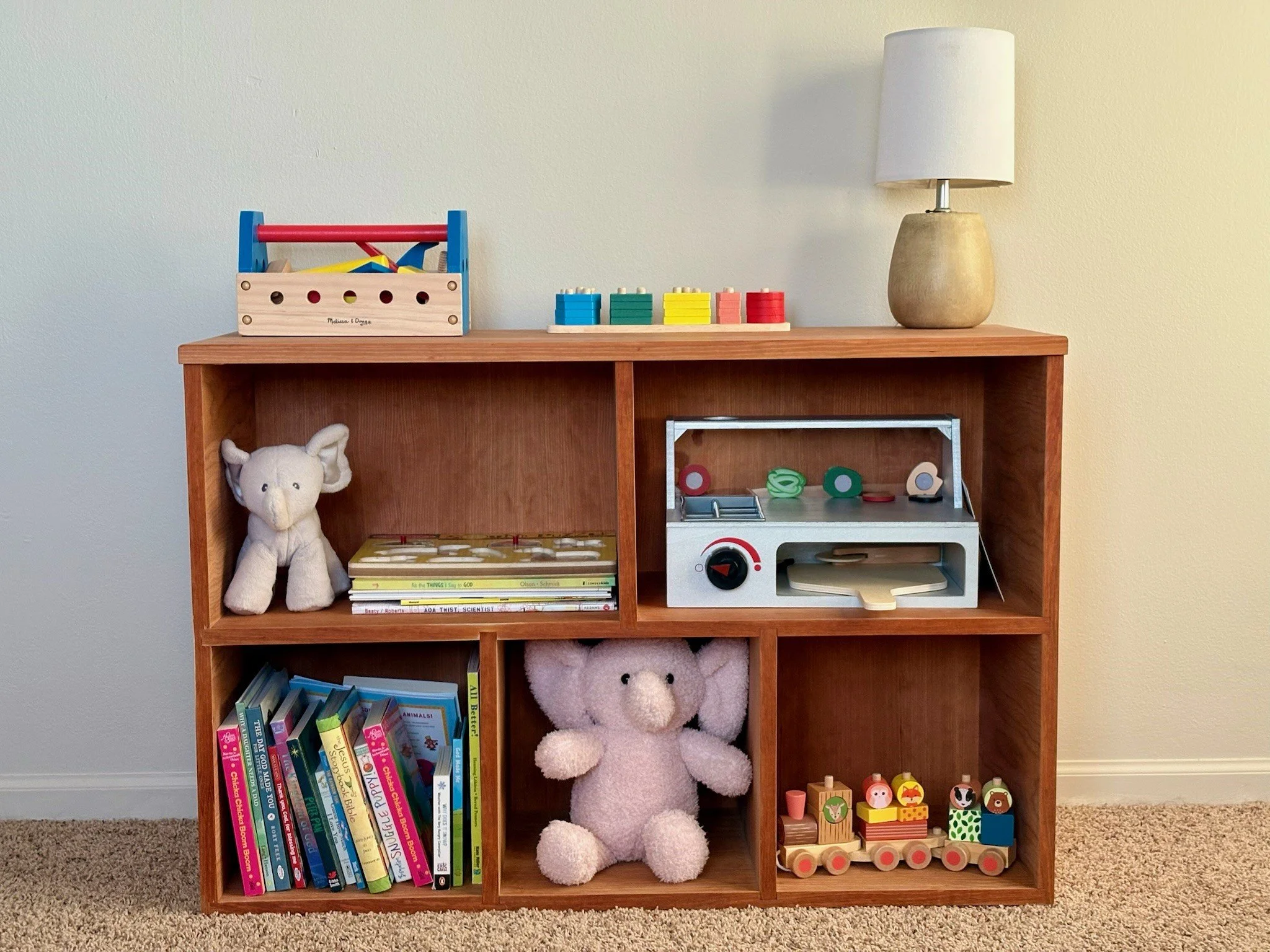 A wooden shelf filled with children's toys, books, and stuffed animals against a plain wall and carpeted floor.