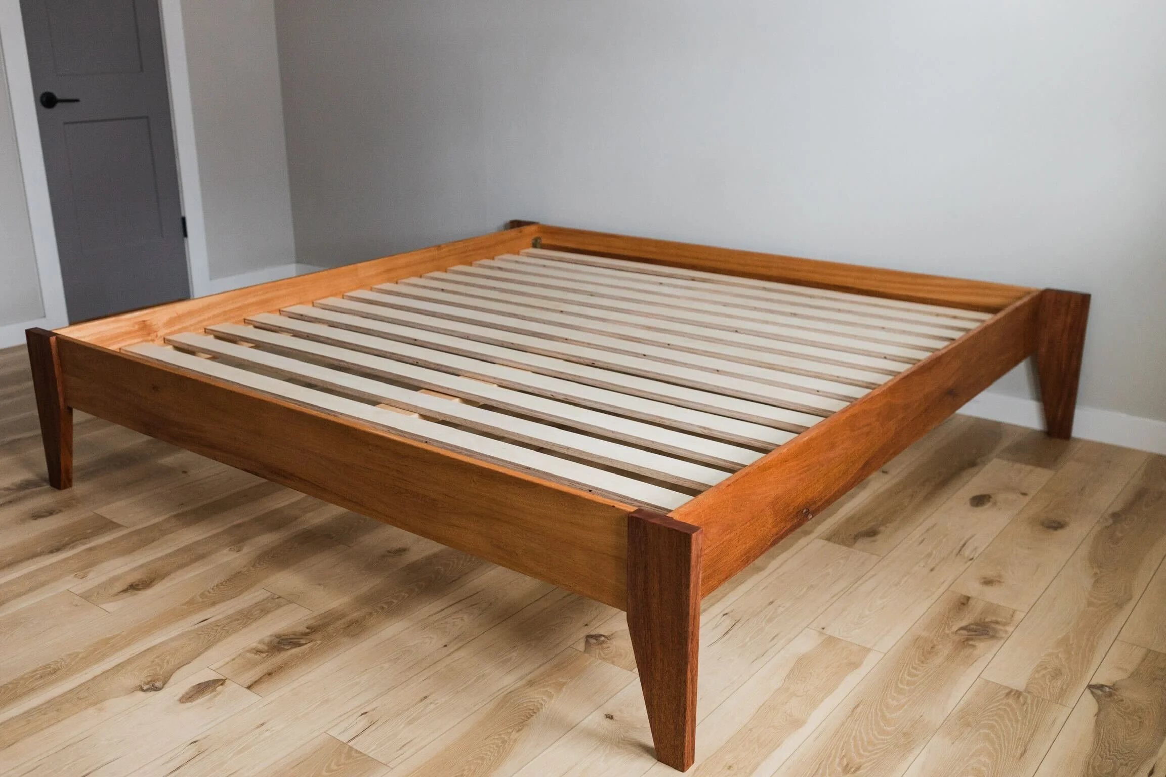 Unassembled wooden bed frame with slats on a light wood floor in a room with gray door and white walls.