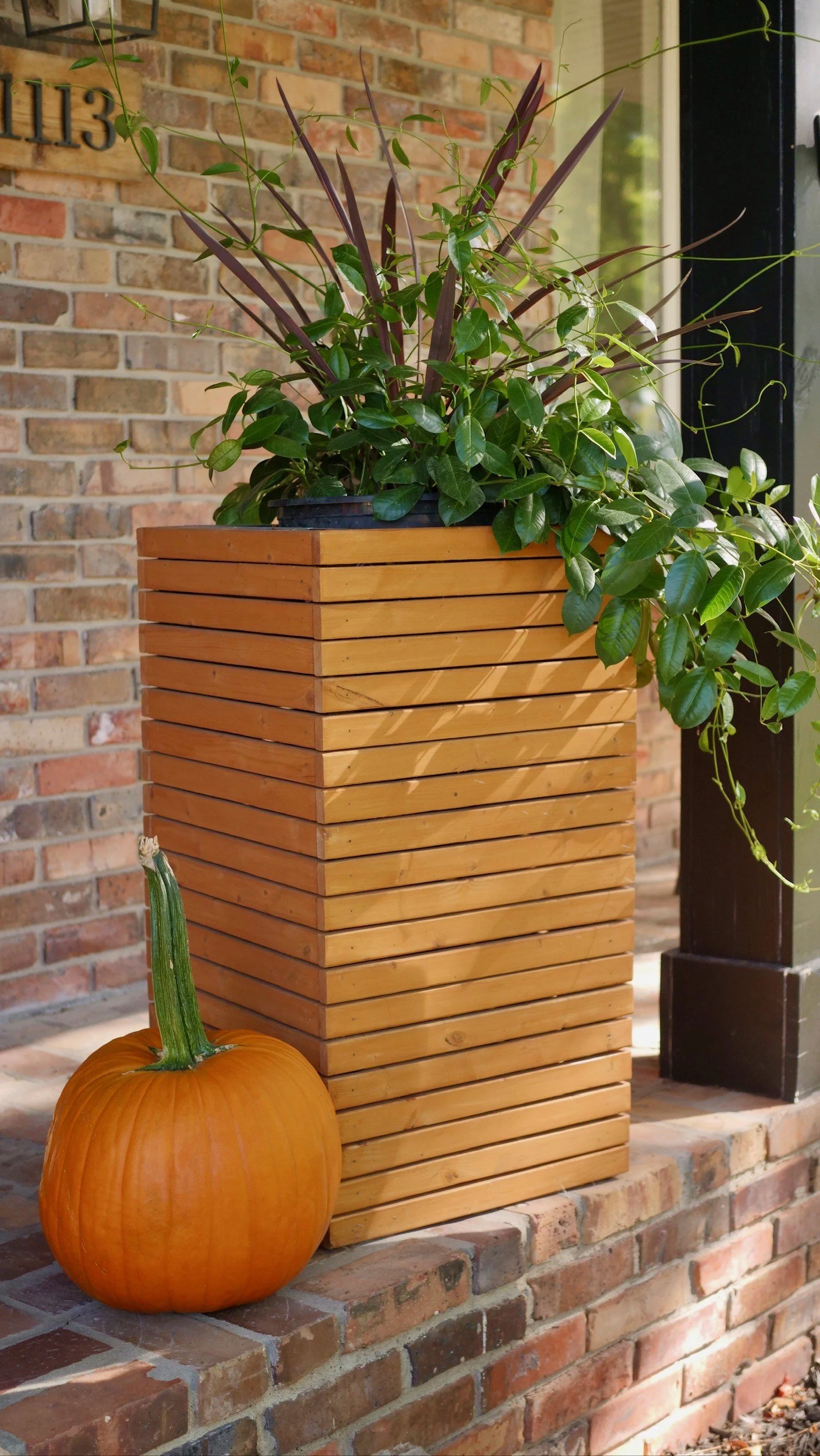 An orange pumpkin and a tall wooden planter box with green plants on a brick porch.