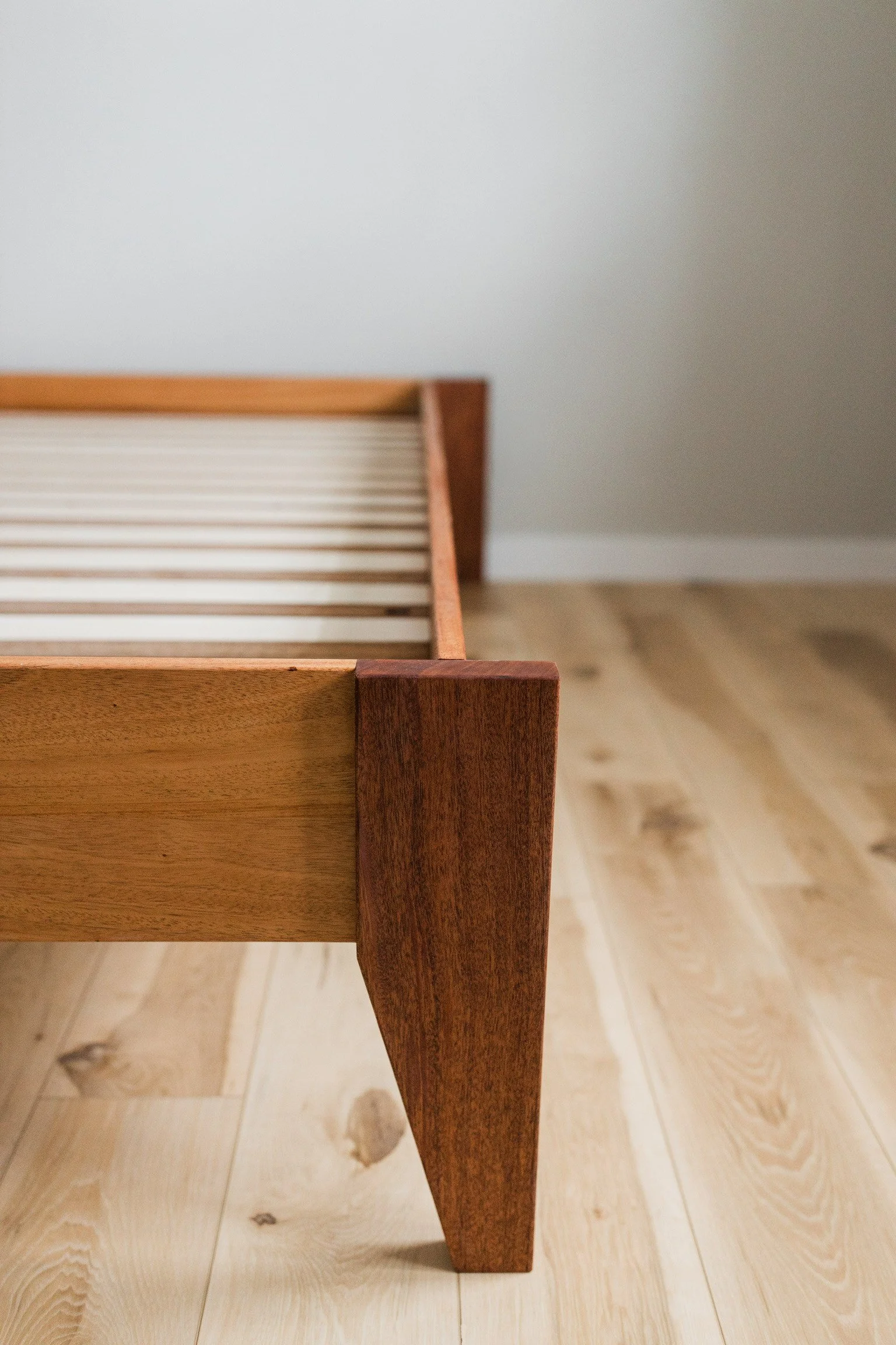 Close-up of a wooden bed frame with a natural finish, showing the corner leg and side, on a light-colored wooden floor, against a plain white wall.