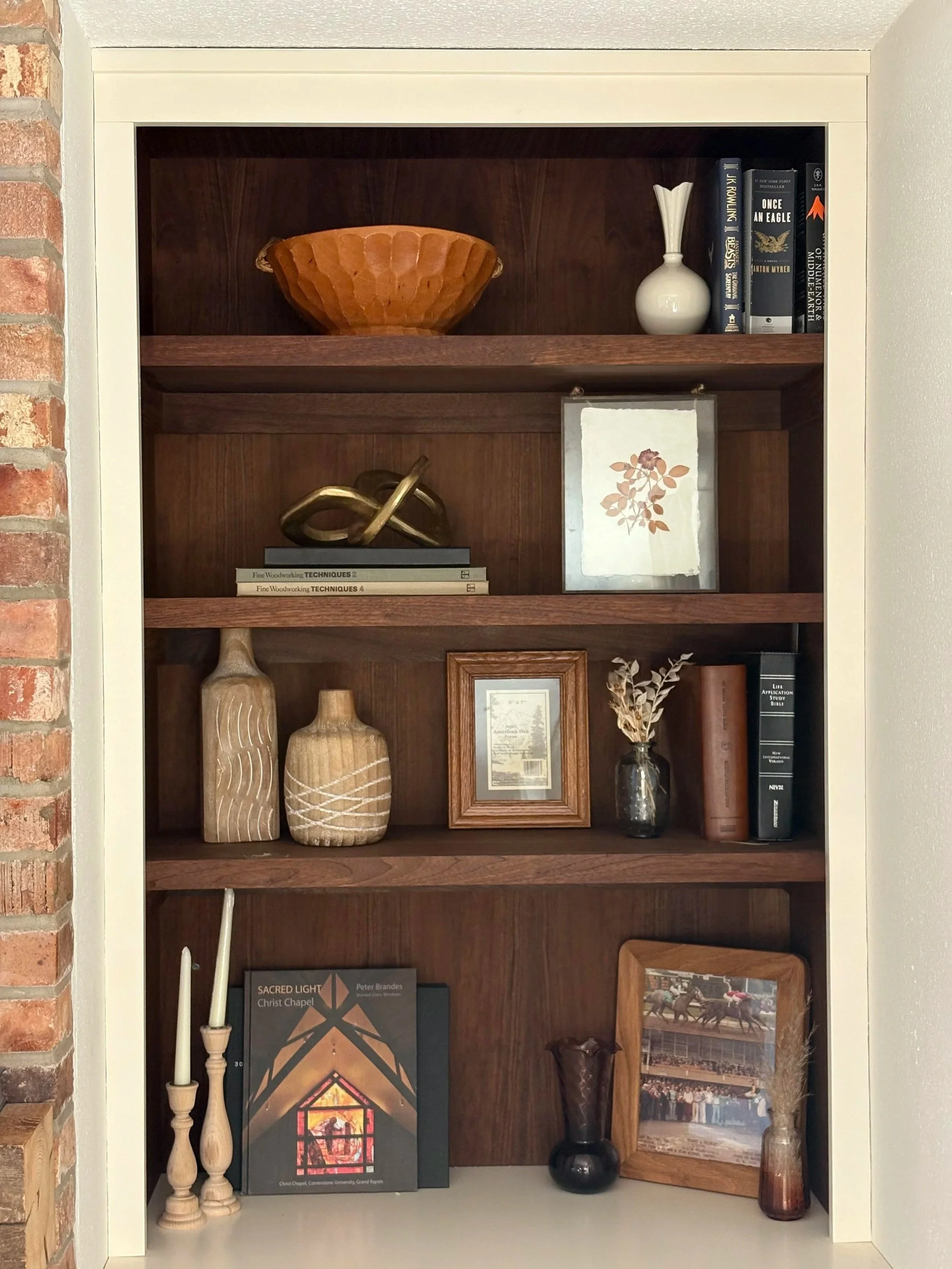 A bookshelf with decorative items, books, and framed pictures. The top shelf has a wooden bowl and a small white vase with a narrow neck. The second shelf has stacked books, a gold sculpture, and a framed artwork. The third shelf has pottery vases, a framed picture, and a small black vase with dried flowers. The bottom shelf has a book about sacred light, two wooden candlesticks with white candles, a framed photo, and two vases.
