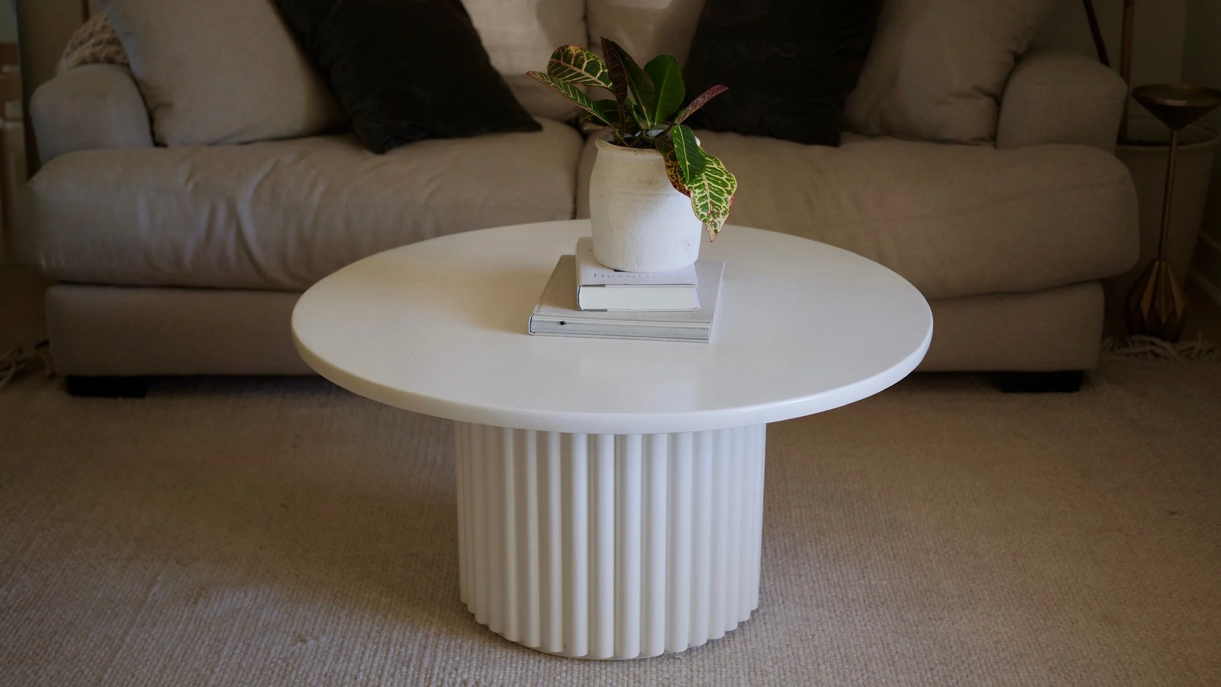 A round white coffee table with a fluted cylindrical base, placed on a beige carpet, with a potted plant, books, and magazines on top. A beige sofa with pillows is in the background.