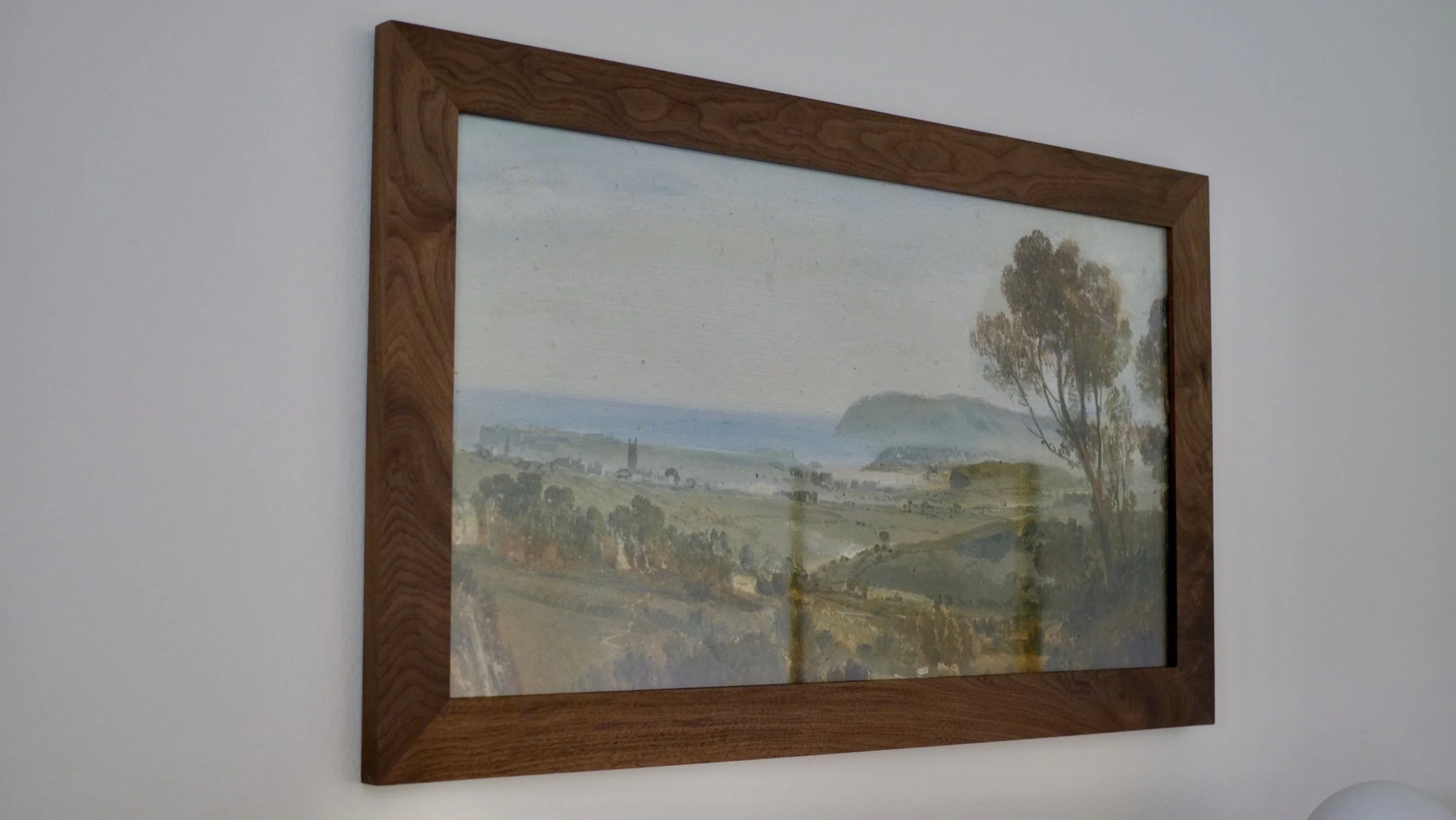 A framed landscape painting depicting trees, rolling hills, and a body of water, hanging on a white wall.