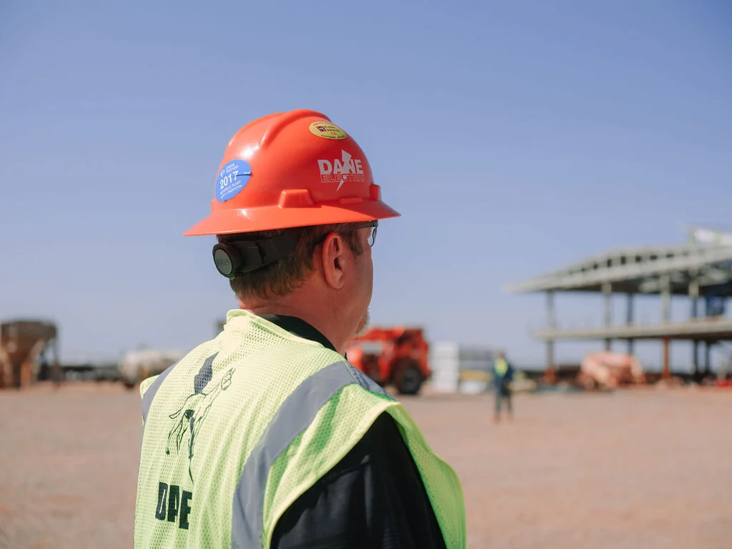 A construction worker wearing an orange safety helmet and a yellow reflective vest stands on a construction site with steel framework and vehicles in the background.