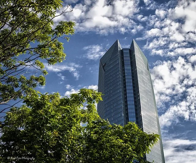 A tall, modern glass skyscraper against a partly cloudy sky, with green trees in the foreground.