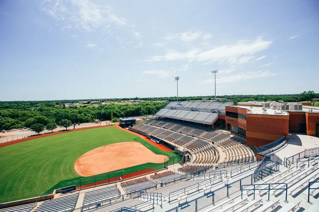Empty baseball stadium with green field and brown dirt infield, surrounded by seating areas and trees in the background under a partly cloudy sky.