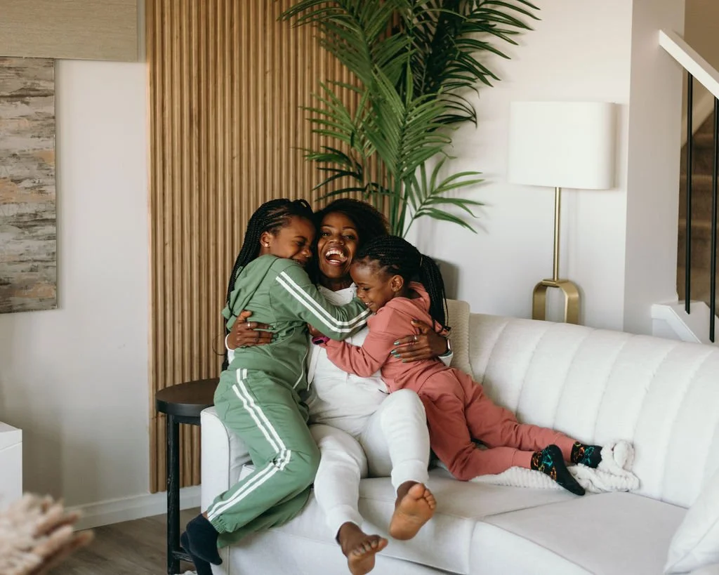 A mom and her two young daughters sitting on a white sofa, hugging and laughing in a well-lit living room with a tall green plant, a modern white lamp, and wooden accents.