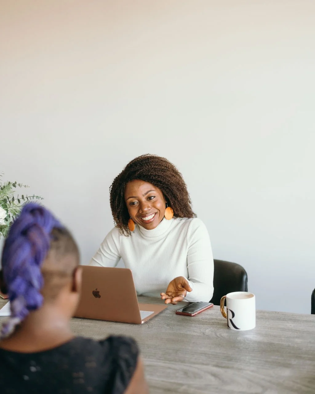 Two women talking in an office setting, one woman with curly hair and wearing white, smiling, while the other woman with purple and shaved hair facing away.