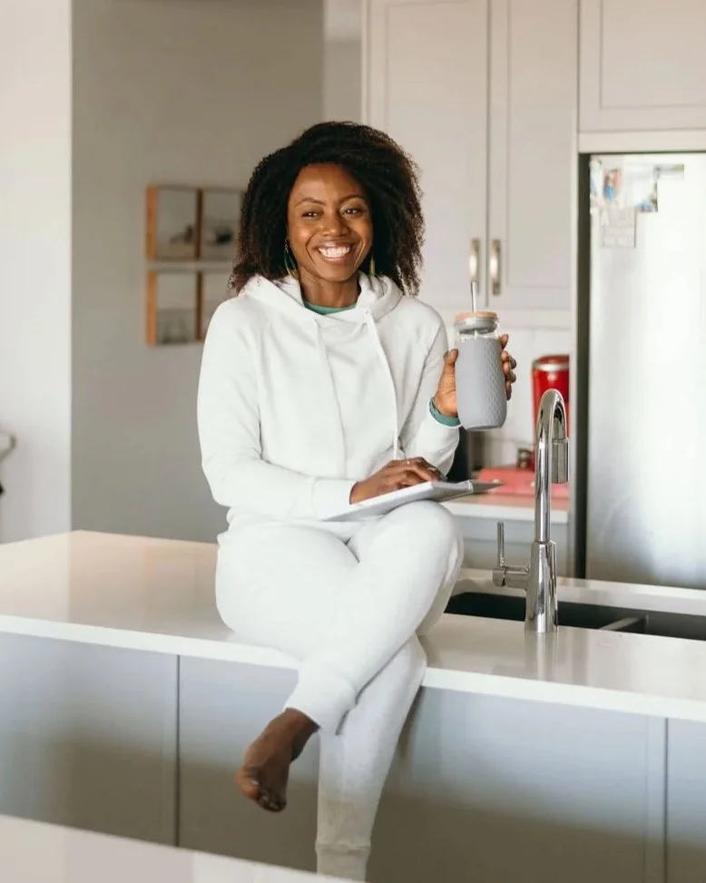 A pleasant smiling woman with curly hair sitting on a kitchen counter, holding a tumbler with a straw, wearing a white hoodie and sweatpants.