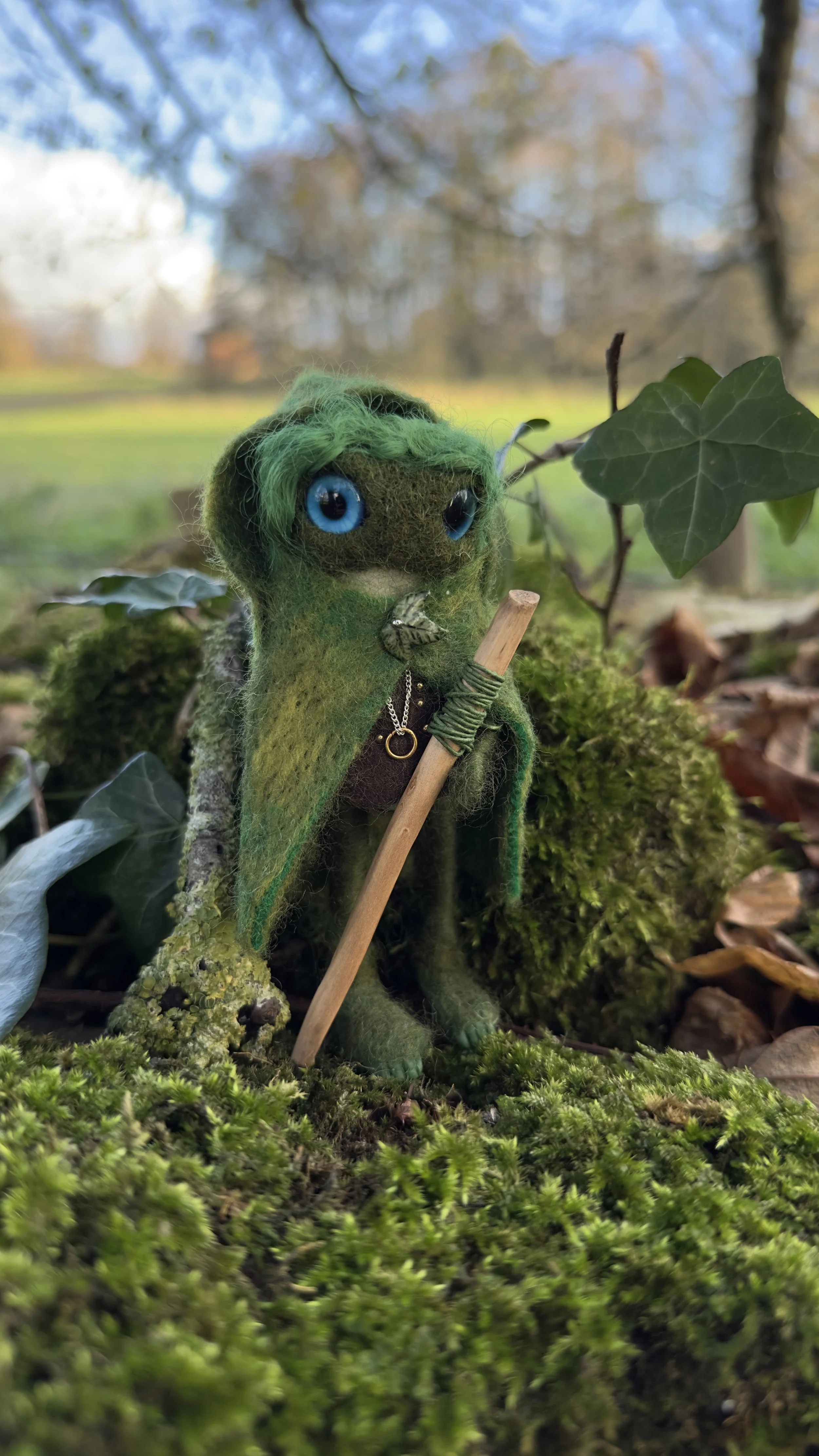 A small felt creature with green hair and big blue eyes dressed as a mystical forest figure, holding a small stick, standing on moss among leaves and plants with trees and cloudy sky in the background.