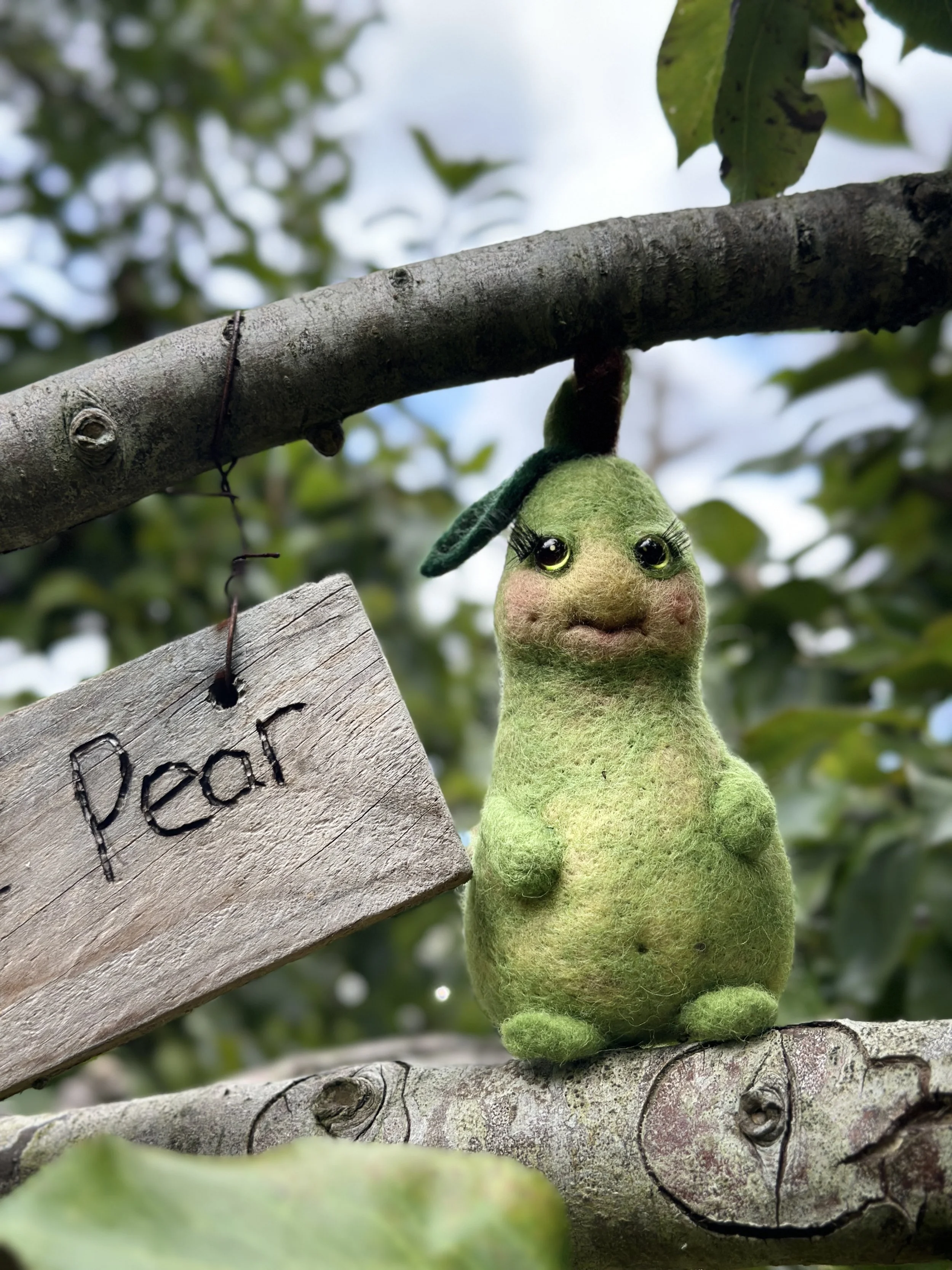 A felted plush toy resembling a green creature with a rounded body, big eyes with long eyelashes, and a small mouth, hanging upside down from a tree branch by its head. A wooden sign with the word 'pearl' written on it is attached to the branch near 