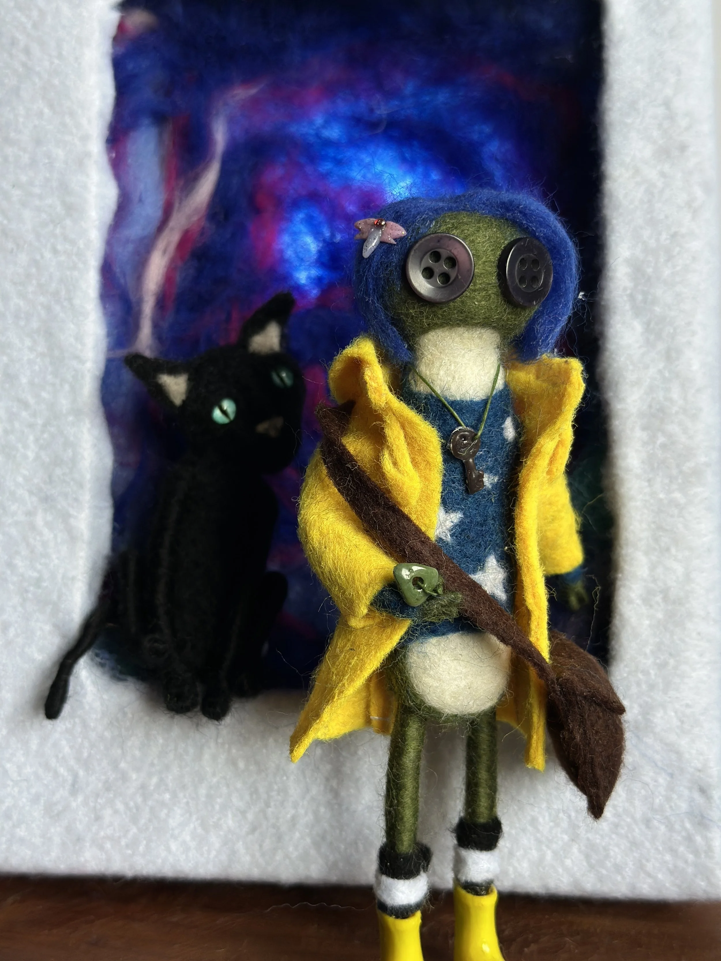 Felted doll resembling a cartoon character with large eyes, blue hair, yellow coat, and green skin, alongside a black felted cat with green eyes, set against a colorful background.