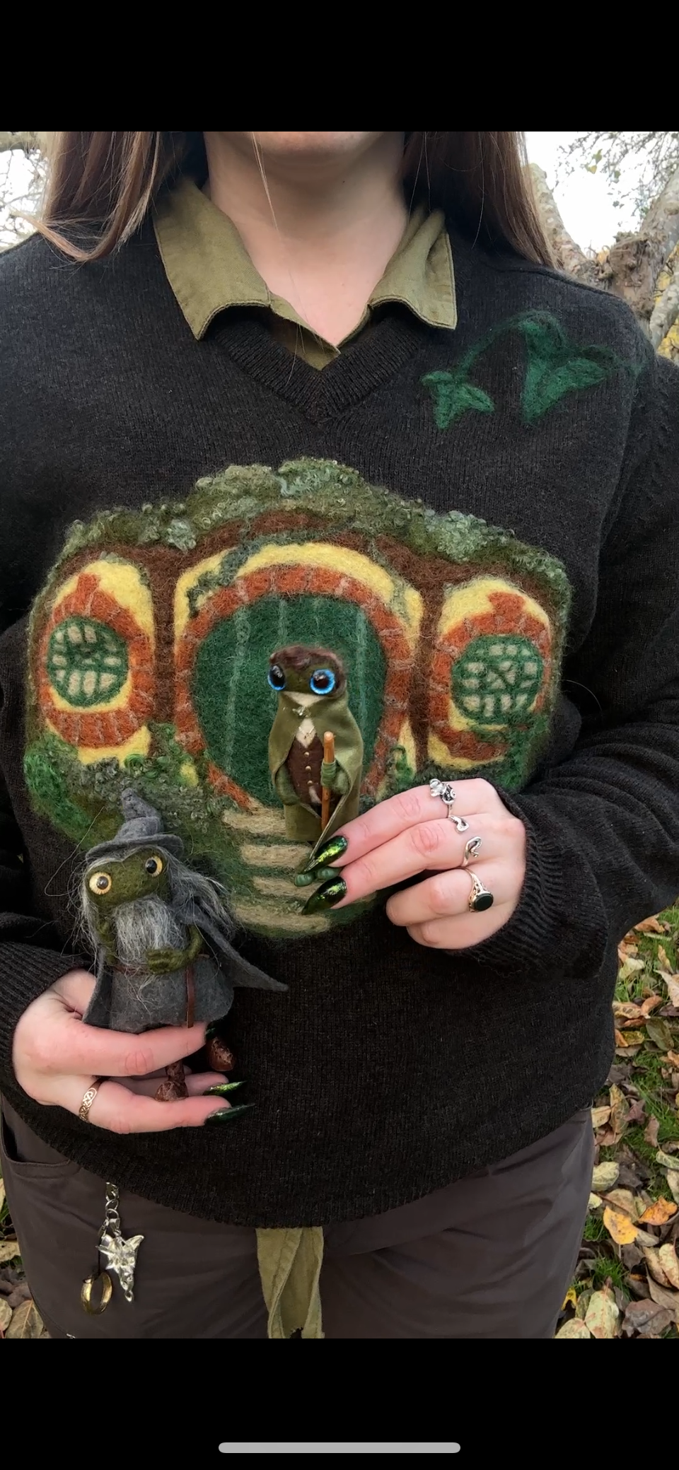 Person wearing a sweater with a Hobbit house design, holding two plush toys of hobbits, one with a walking stick, outdoors with fallen leaves.