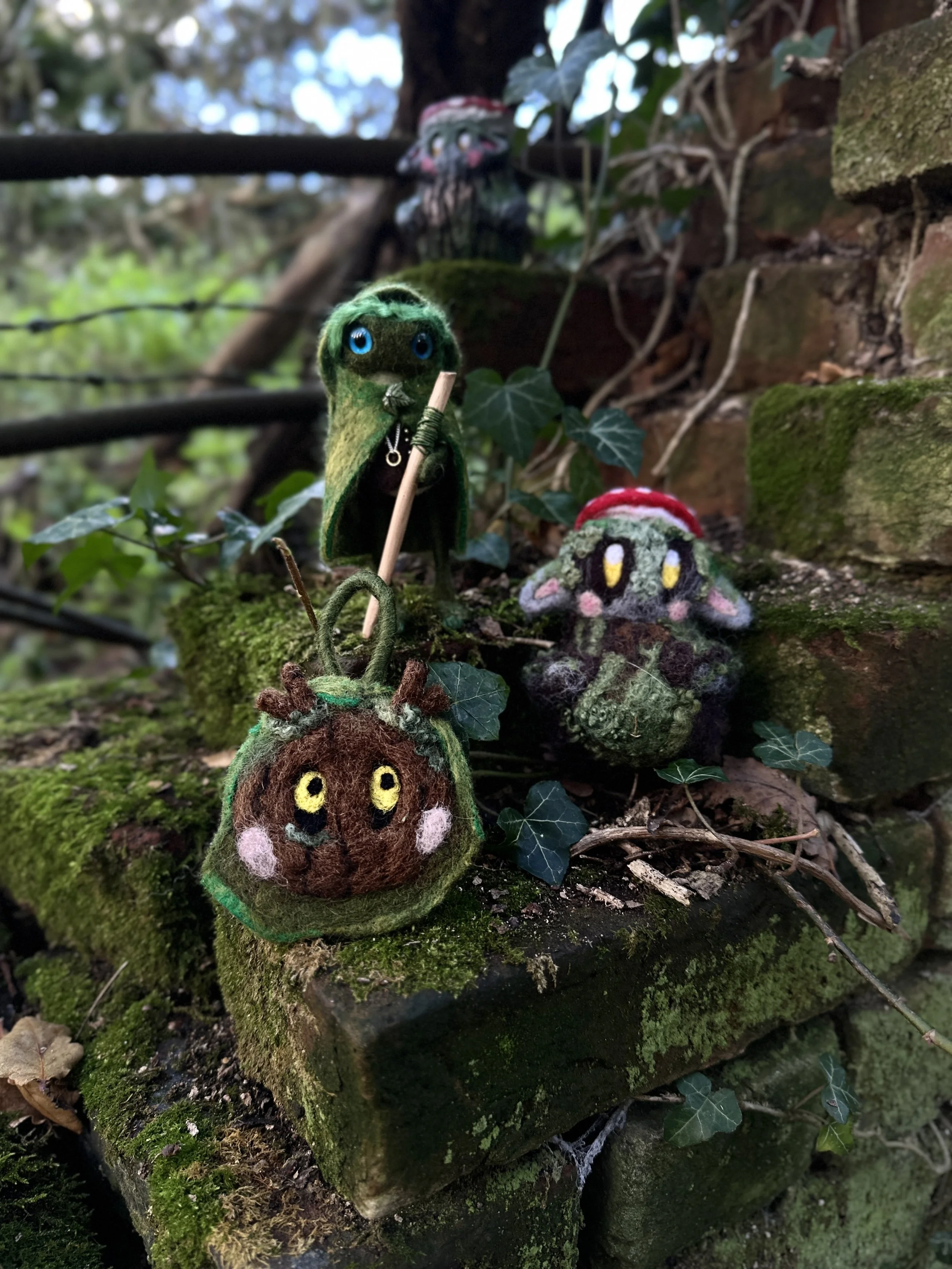 Three handmade felt creatures resembling fantasy characters are placed on a moss-covered brick ledge outdoors, with one figure holding a staff and wearing a cloak, while the other two have large eyes and are adorned with Christmas hats.