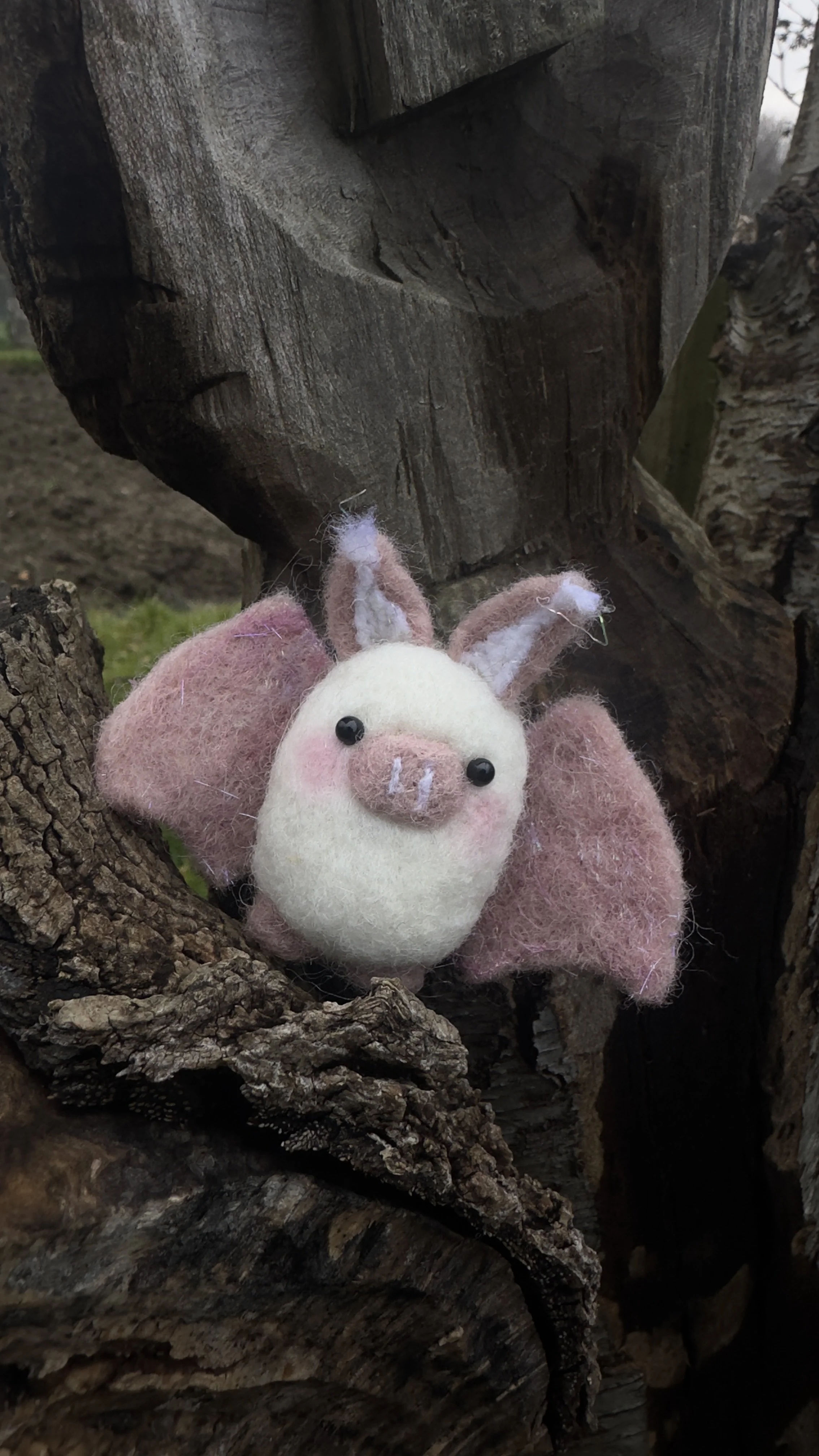 A handmade felted pig with bunny ears and pink and white colors, placed on a piece of firewood outdoors.