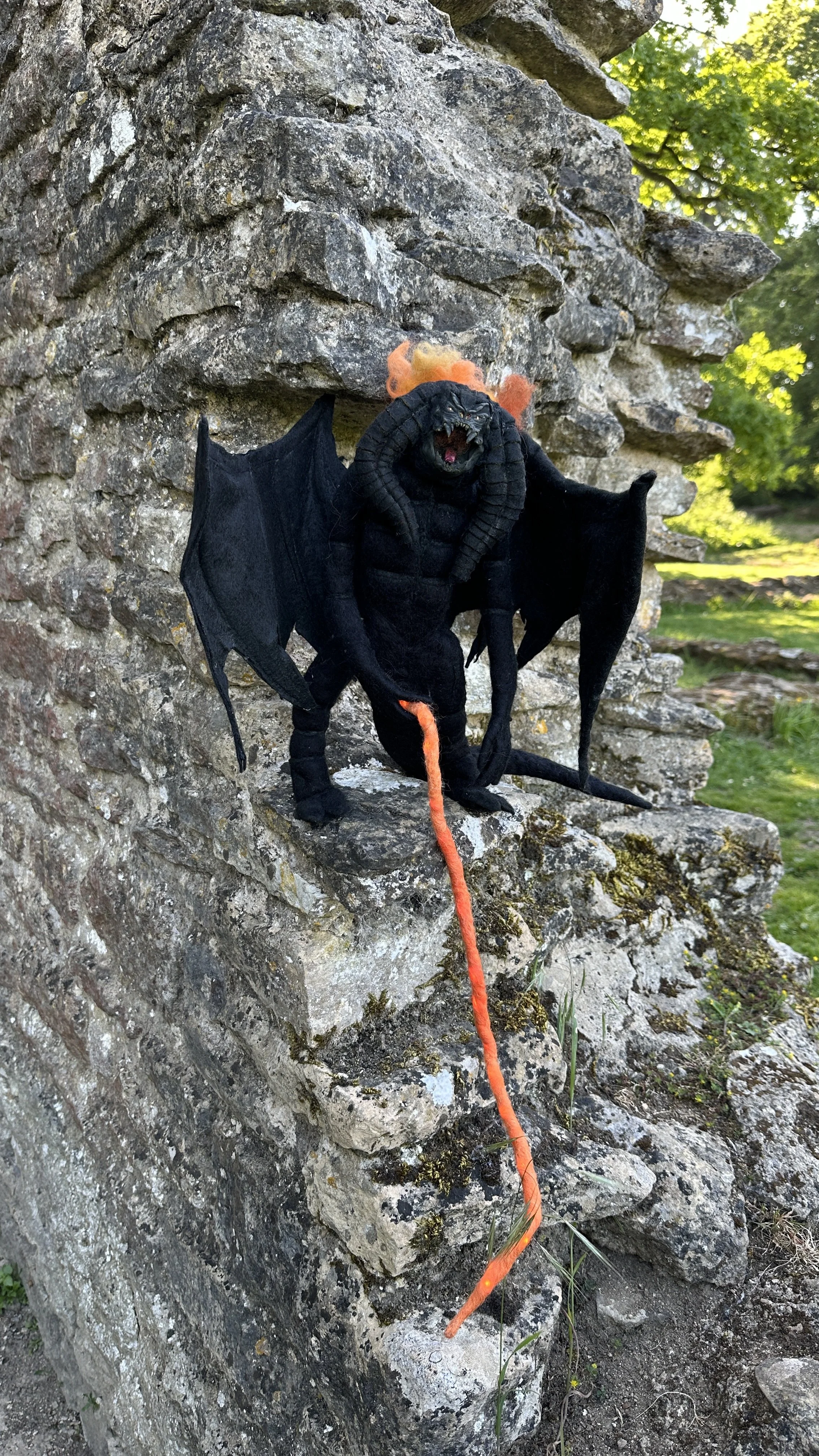 A Halloween decoration of a dragon sitting on a stone ledge outdoors, surrounded by trees and grass. The dragon is black with orange accents, has wings, horns, and a fierce expression.