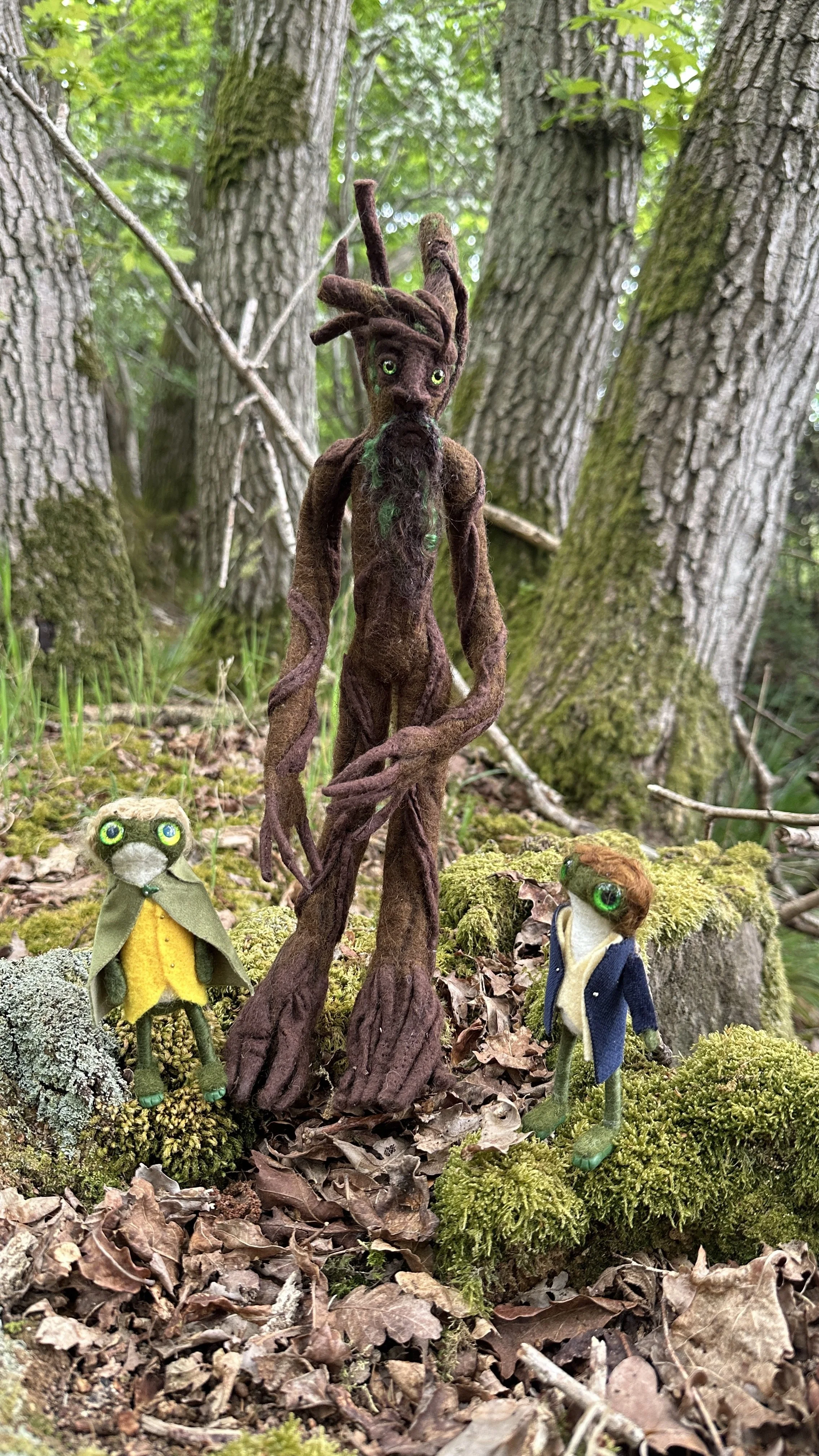 Three handmade dolls with large green eyes in a forest setting with trees, moss, and leaves. One doll has brown dreadlocks, standing in the center, with a beard and dressed in earthy tones. The other two dolls are smaller, one with blonde hair and a 