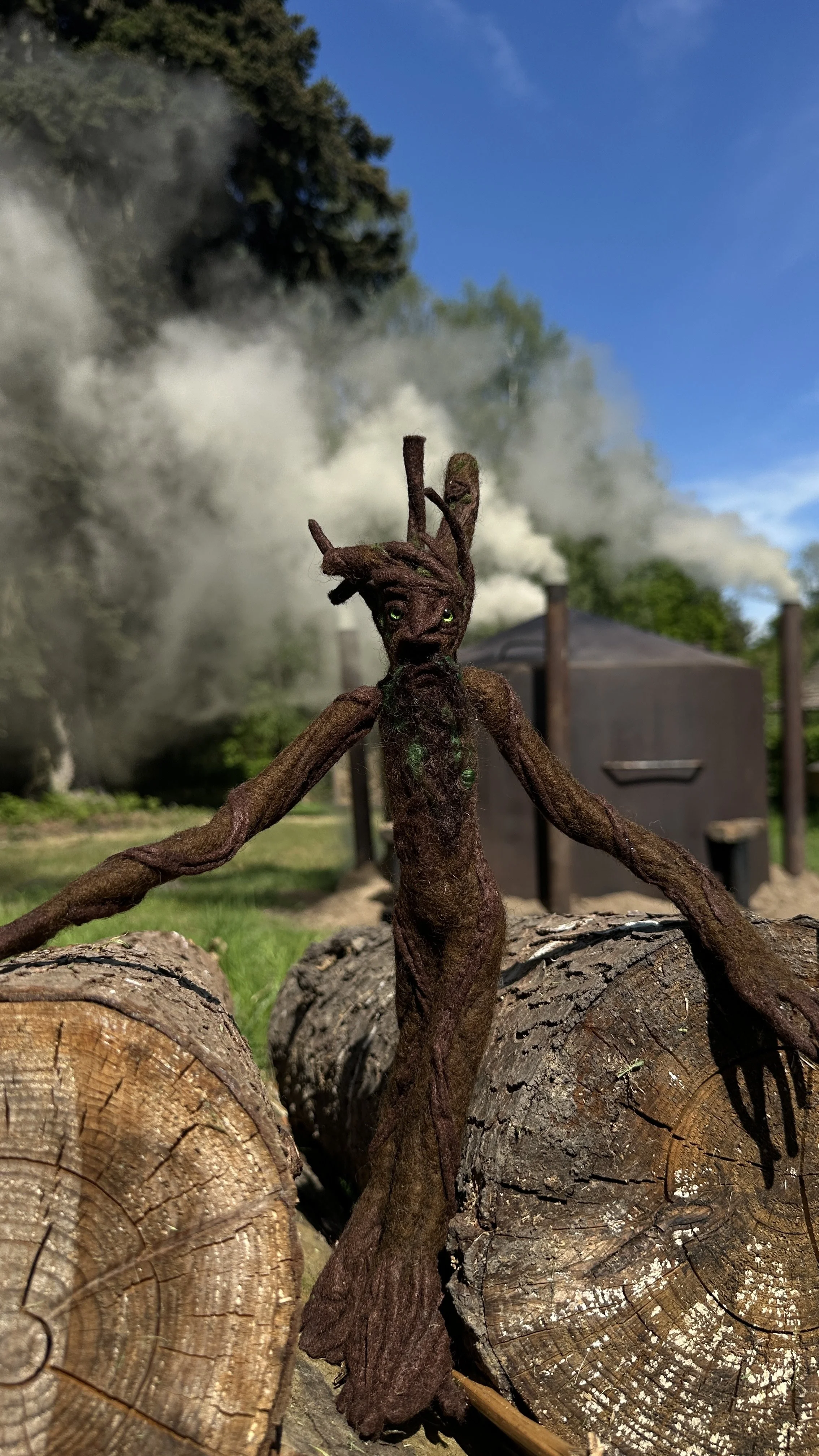 A doll made to look like a tree or forest spirit stands on logs outdoors. In the background, there is a wooden structure and smoke or steam rising into the blue sky.