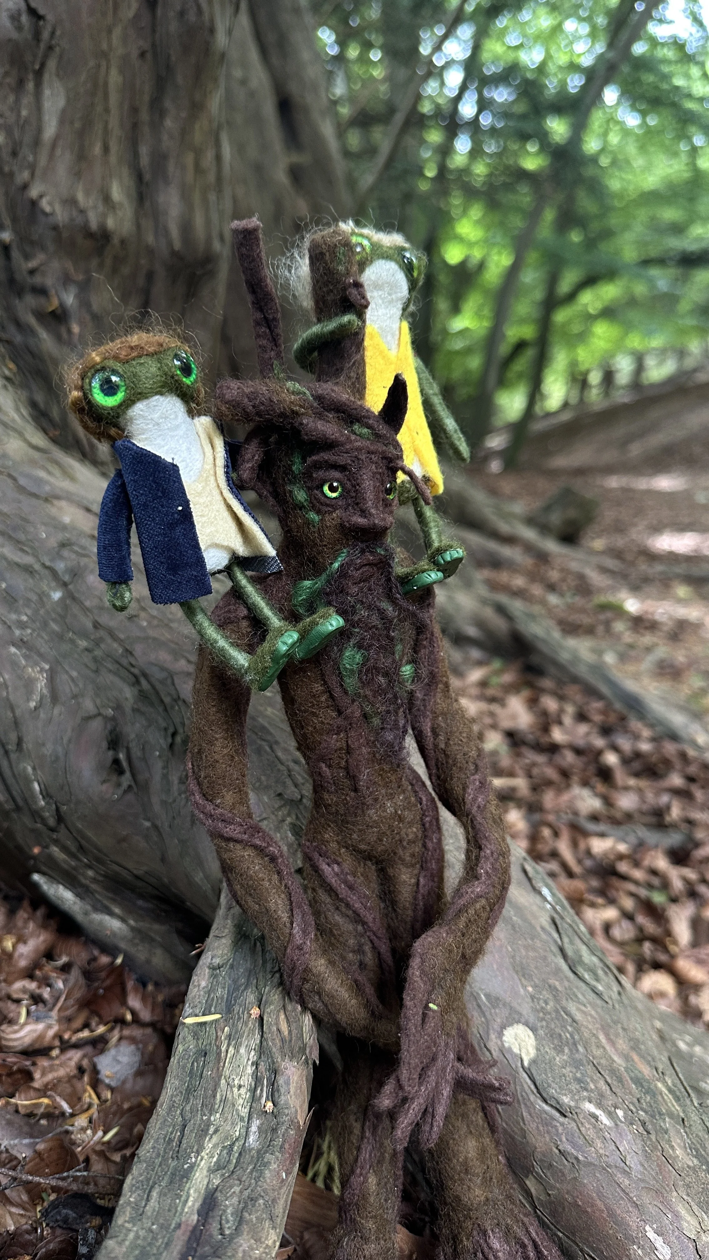 Felted animal dolls resembling mythical creatures or forest spirits, sitting on a fallen tree in a forest. The creatures have green eyes, pointed ears, and are dressed in colorful garments. They have expressive faces and are surrounded by greenery.