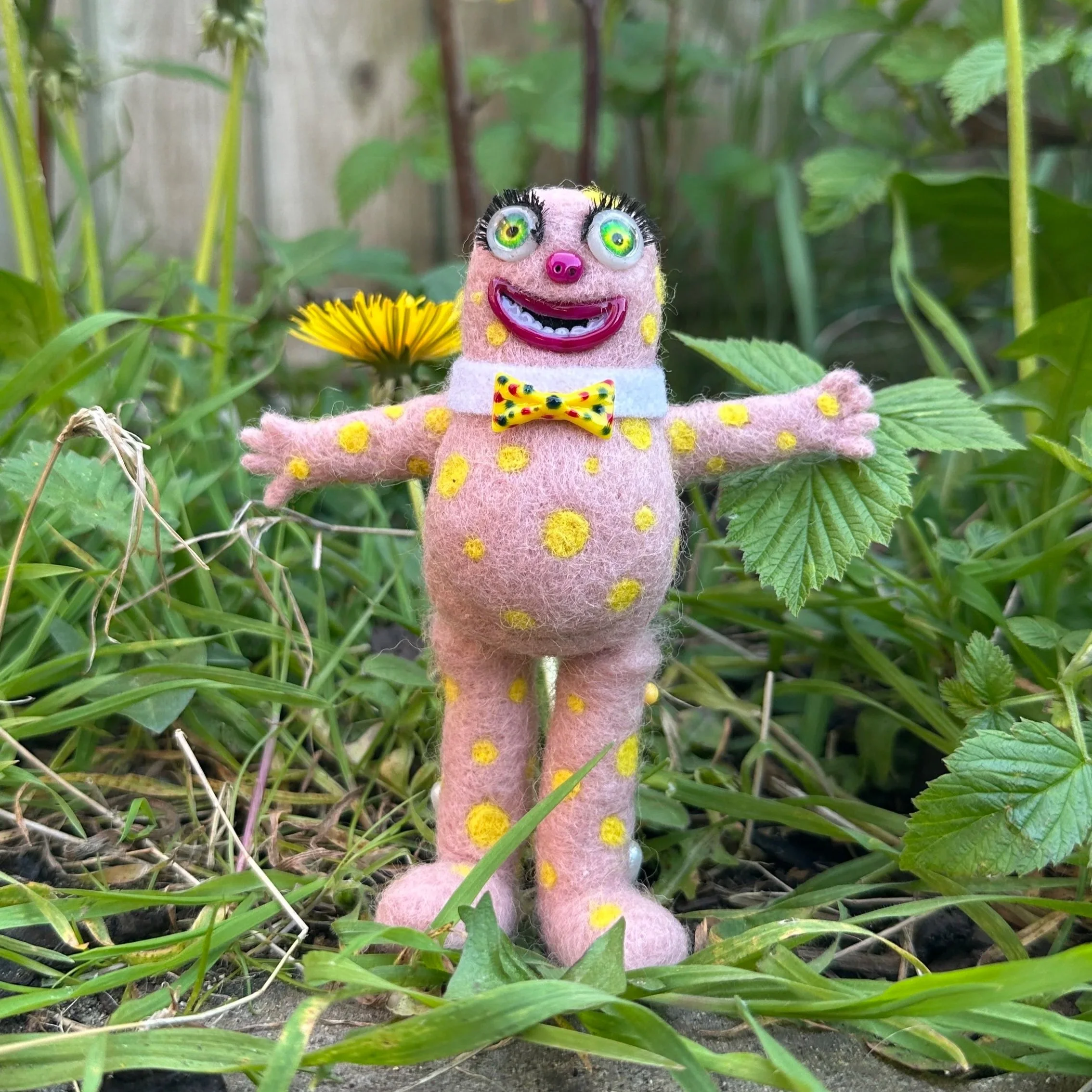 A colorful felt doll with exaggerated facial features, yellow polka dots, a yellow bow tie, and outstretched arms, standing among green plants and a yellow flower.