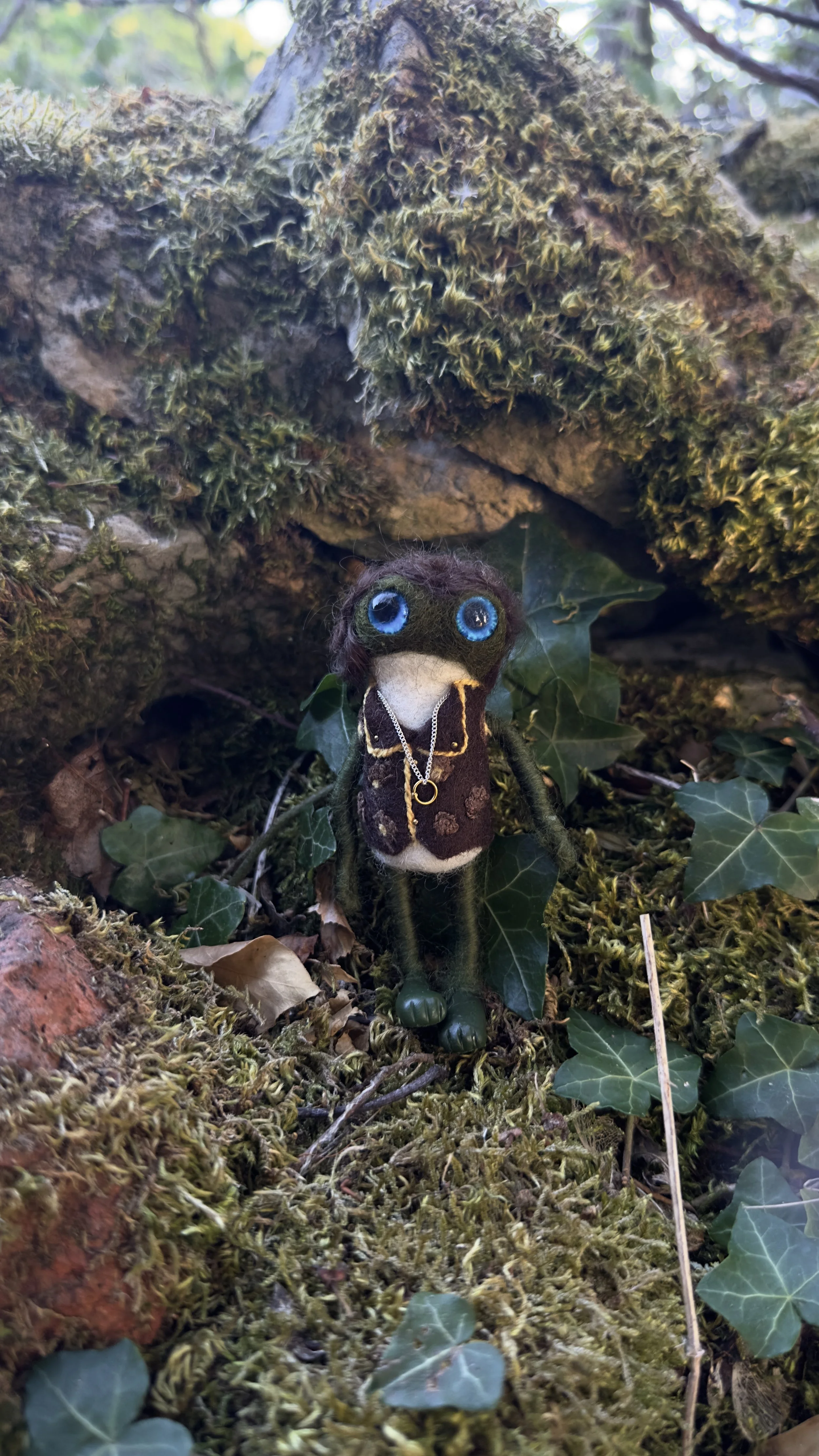 A felted doll with big blue eyes, dark brown hair, and green limbs, dressed in a brown vest and necklace, standing among moss and ivy on forest ground with a tree root overhead.