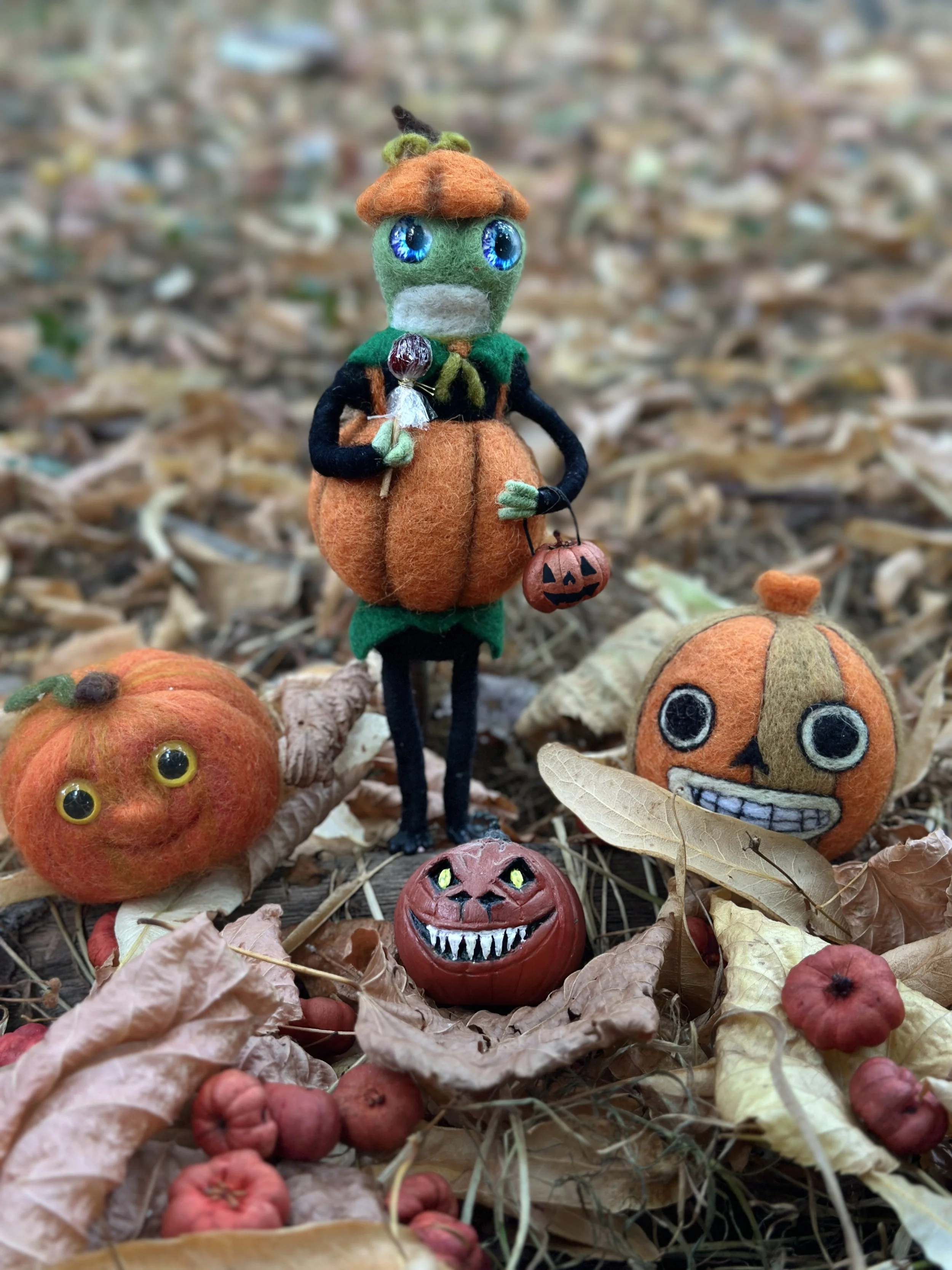 Felt Halloween scene with a doll dressed as a scarecrow holding a lollipop and a small pumpkin bucket, surrounded by felt mini pumpkins and dry autumn leaves.