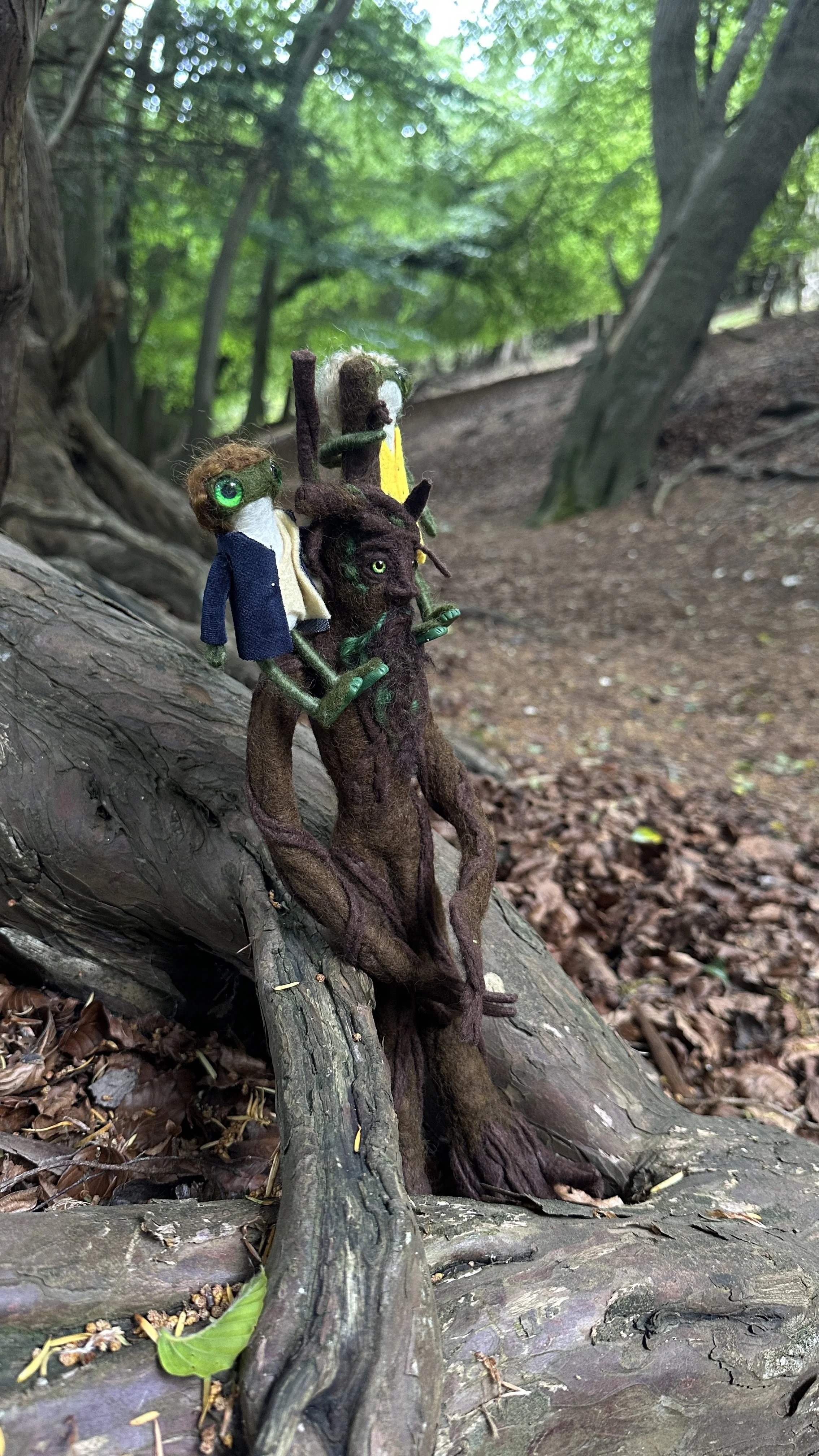 Felted dolls resembling characters from 'Fantastic Beasts' are perched on a fallen tree in a forest, with green foliage and a dirt trail in the background.