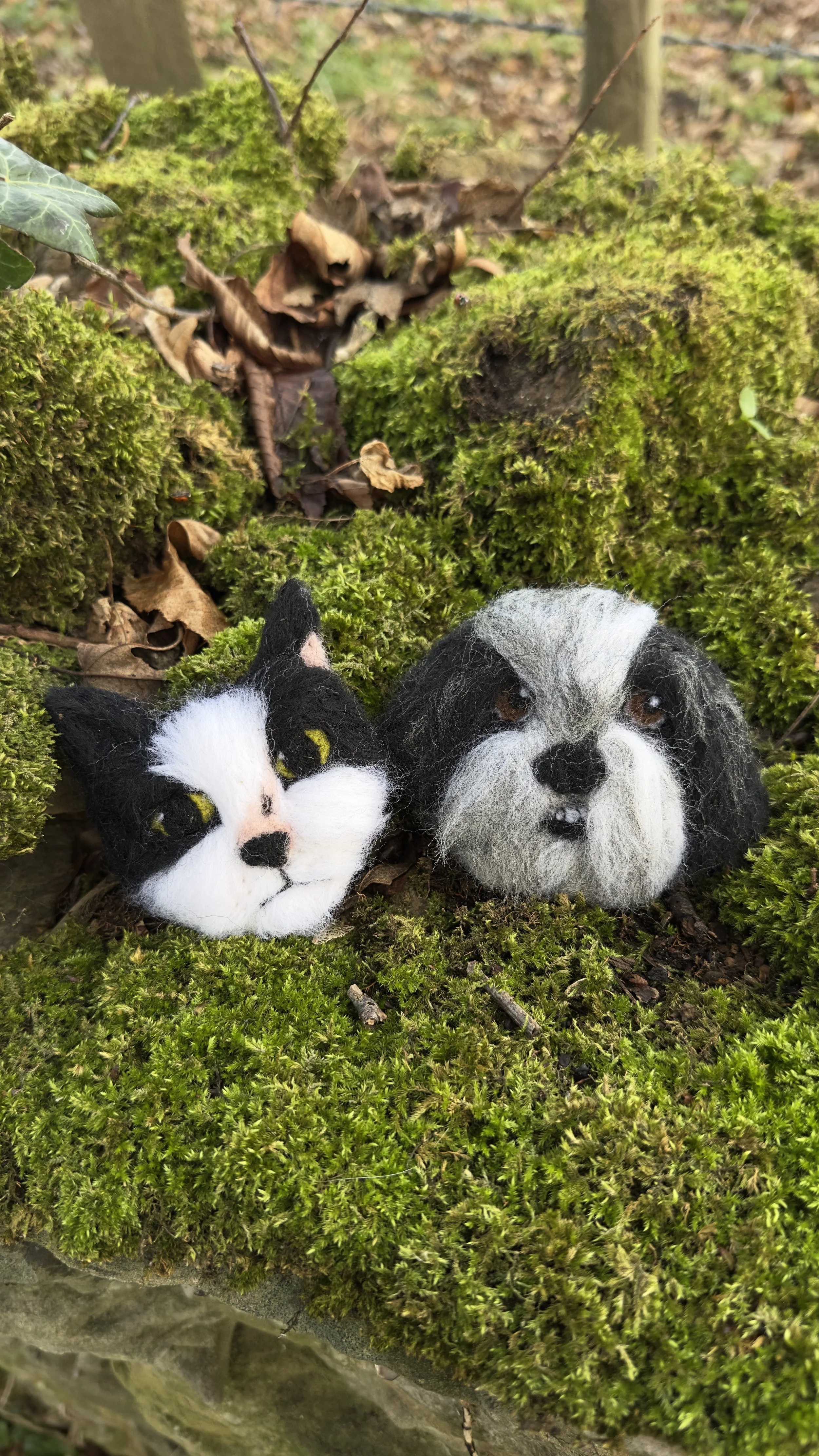 Two felted animal heads, a cat and a dog, placed among moss and forest ground leaves outdoors.