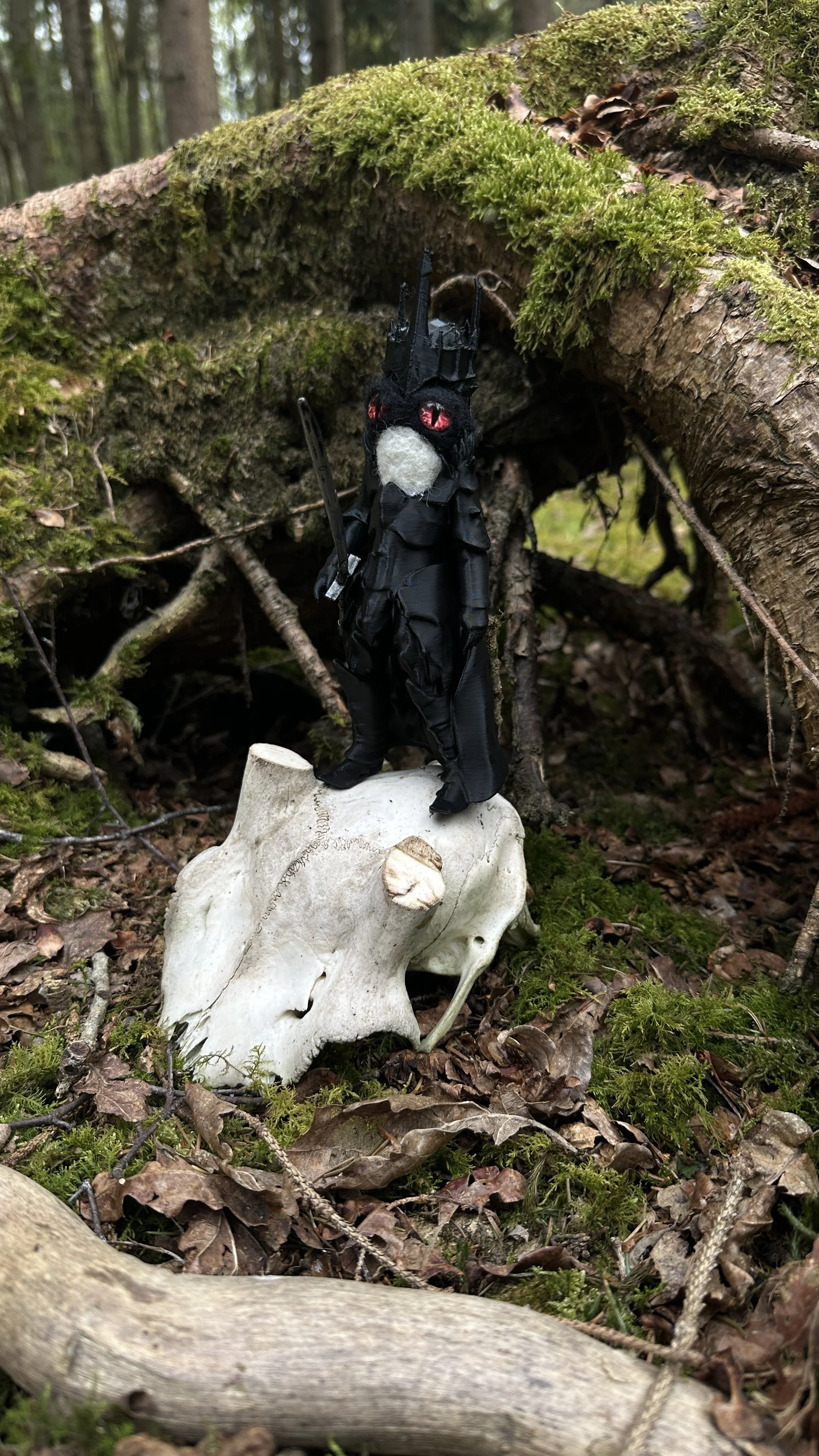 A small, black, fantasy creature figurine with red eyes and a white nose, dressed in a black cloak and holding a sword, standing on top of a human skull in a forest setting with moss-covered logs and branches in the background.
