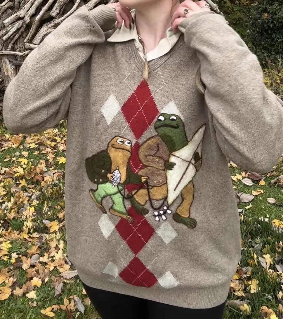 A woman is wearing a beige sweater with an argyle pattern and cartoon frog characters holding fishing gear. She is outdoors among fallen autumn leaves.