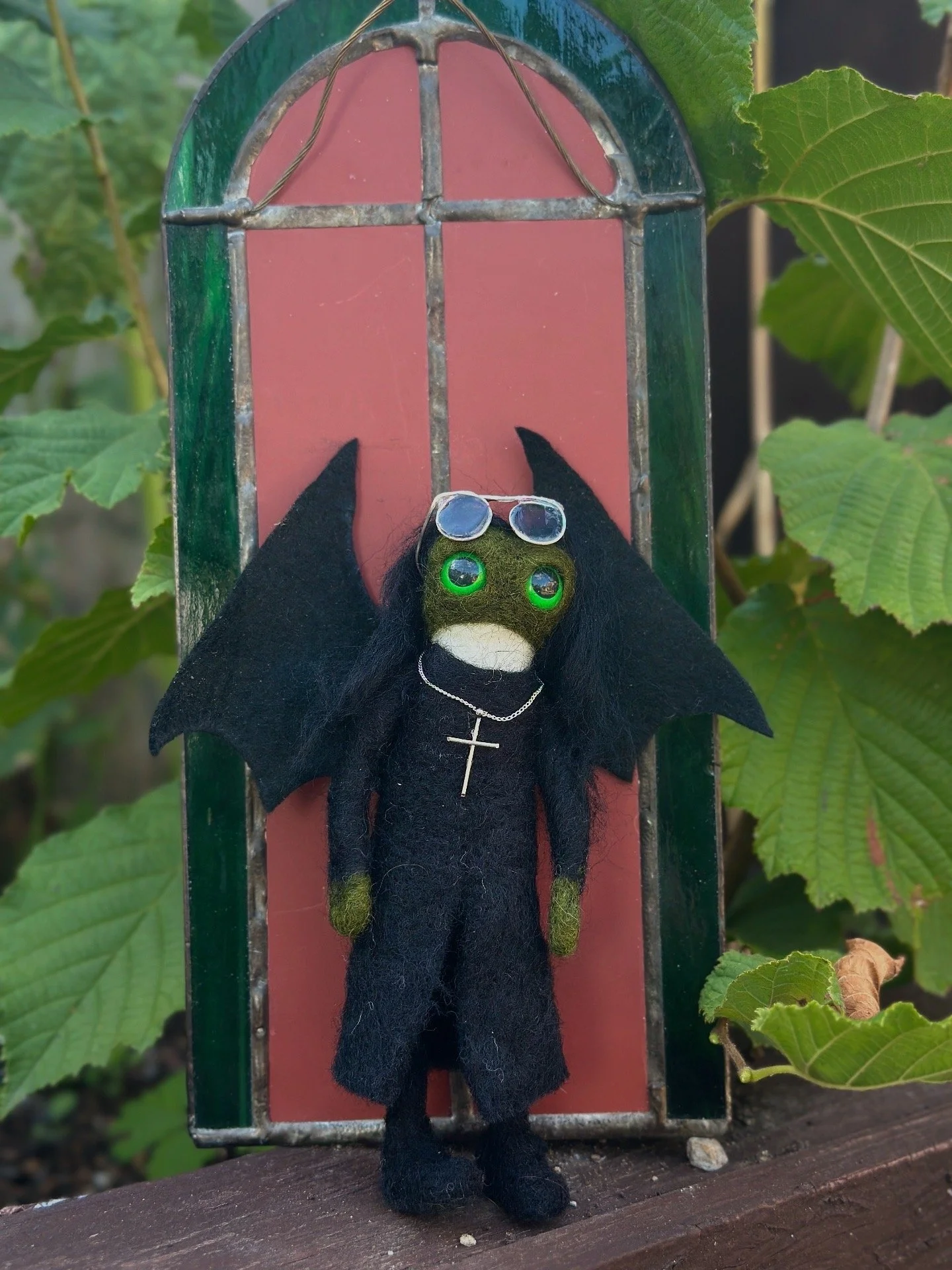 A handmade doll with green eyes, wearing black clothes, a cross necklace, and sunglasses, standing in front of a stained glass window with pink and green sections, surrounded by green leaves.