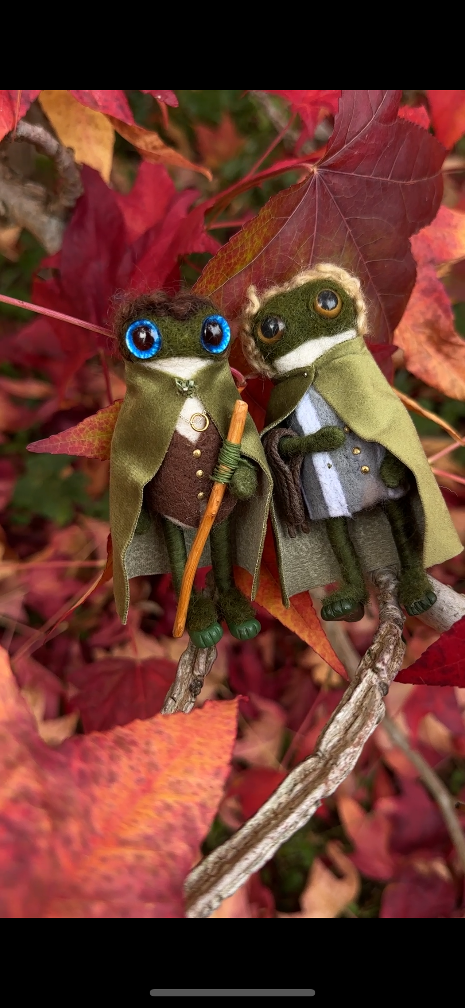 Two felted frog dolls dressed as hobbits, with green cloaks, one holding a staff, against a backdrop of colorful autumn leaves.