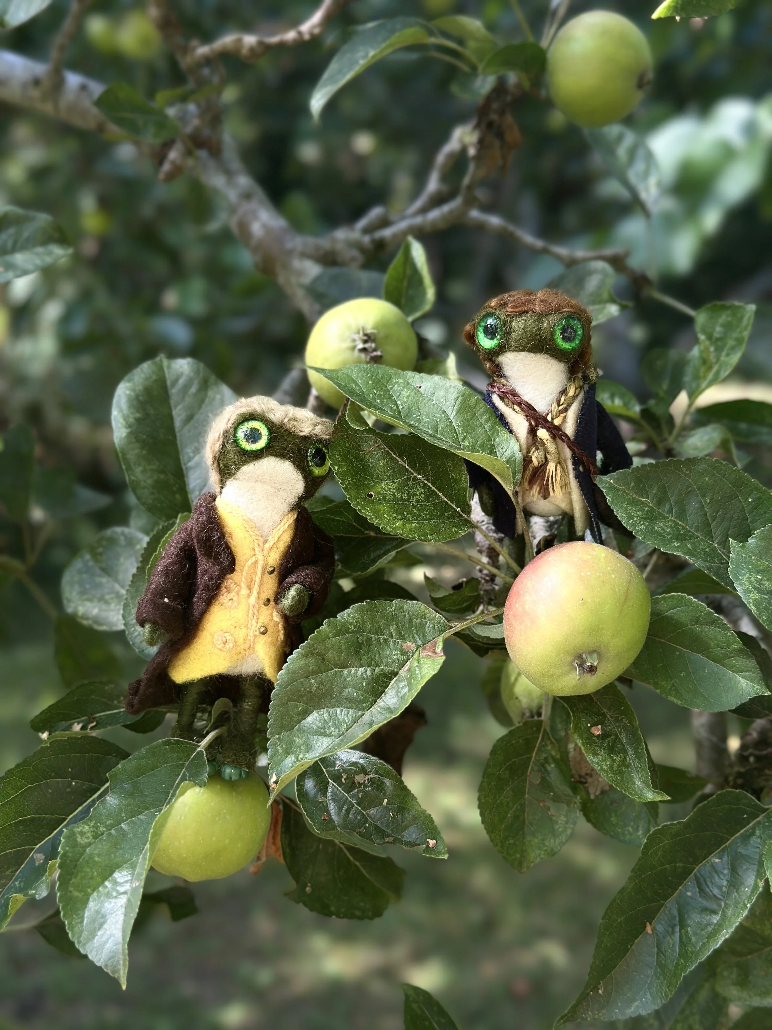 Two felted animal figures resembling frogs, dressed in clothing, sitting on a branch with apple-like fruits and green leaves.