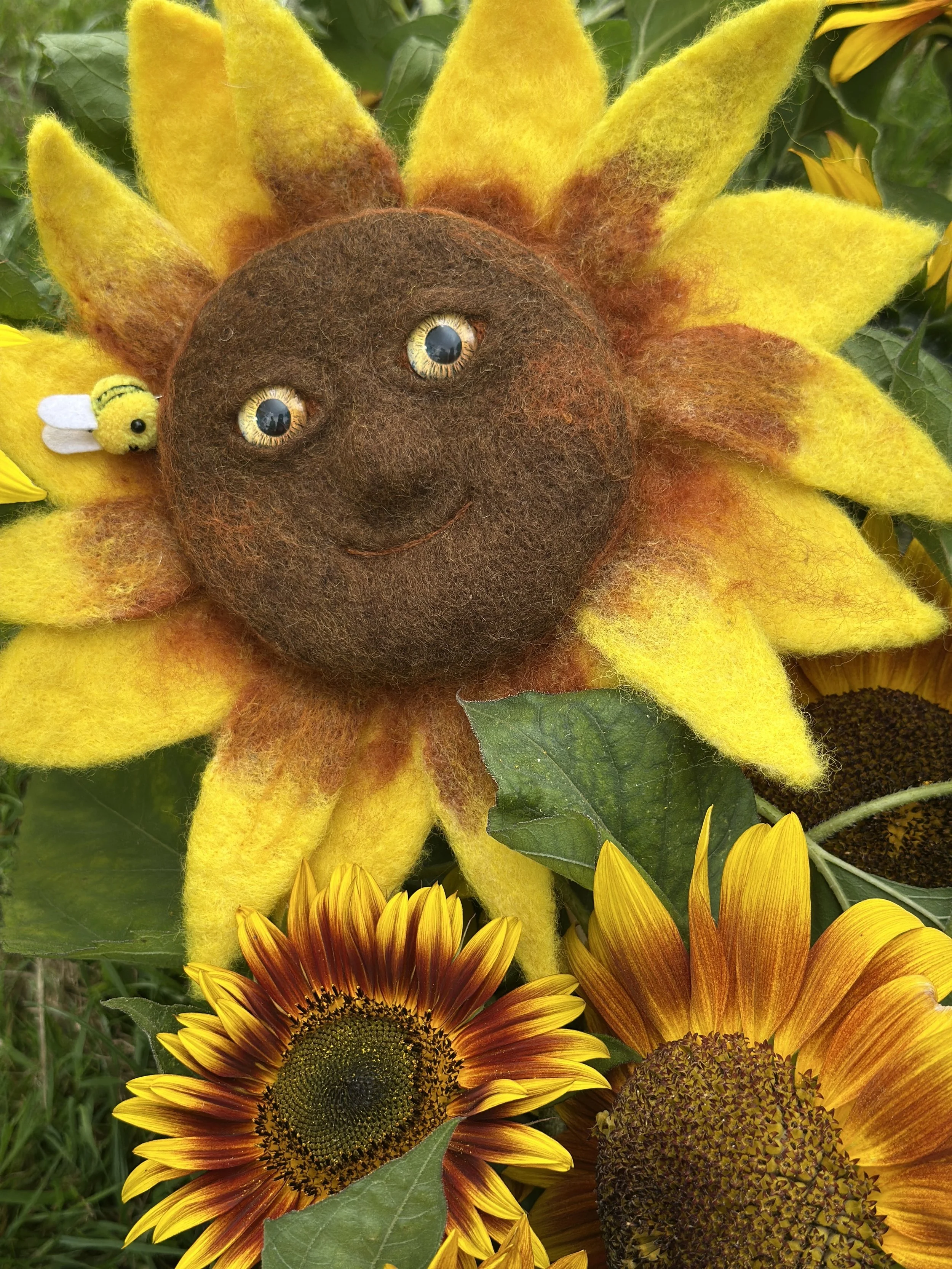 A felted sunflower with a smiling face, featuring a brown center and yellow petals, surrounded by real sunflowers and green leaves, with a small plush bee attached to the sunflower.