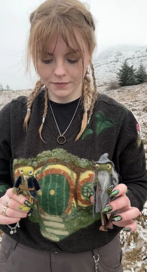 A young woman with red hair in braids holds two frog felted dolls, standing outdoors on a cloudy, snowy mountain with evergreen trees in the background.