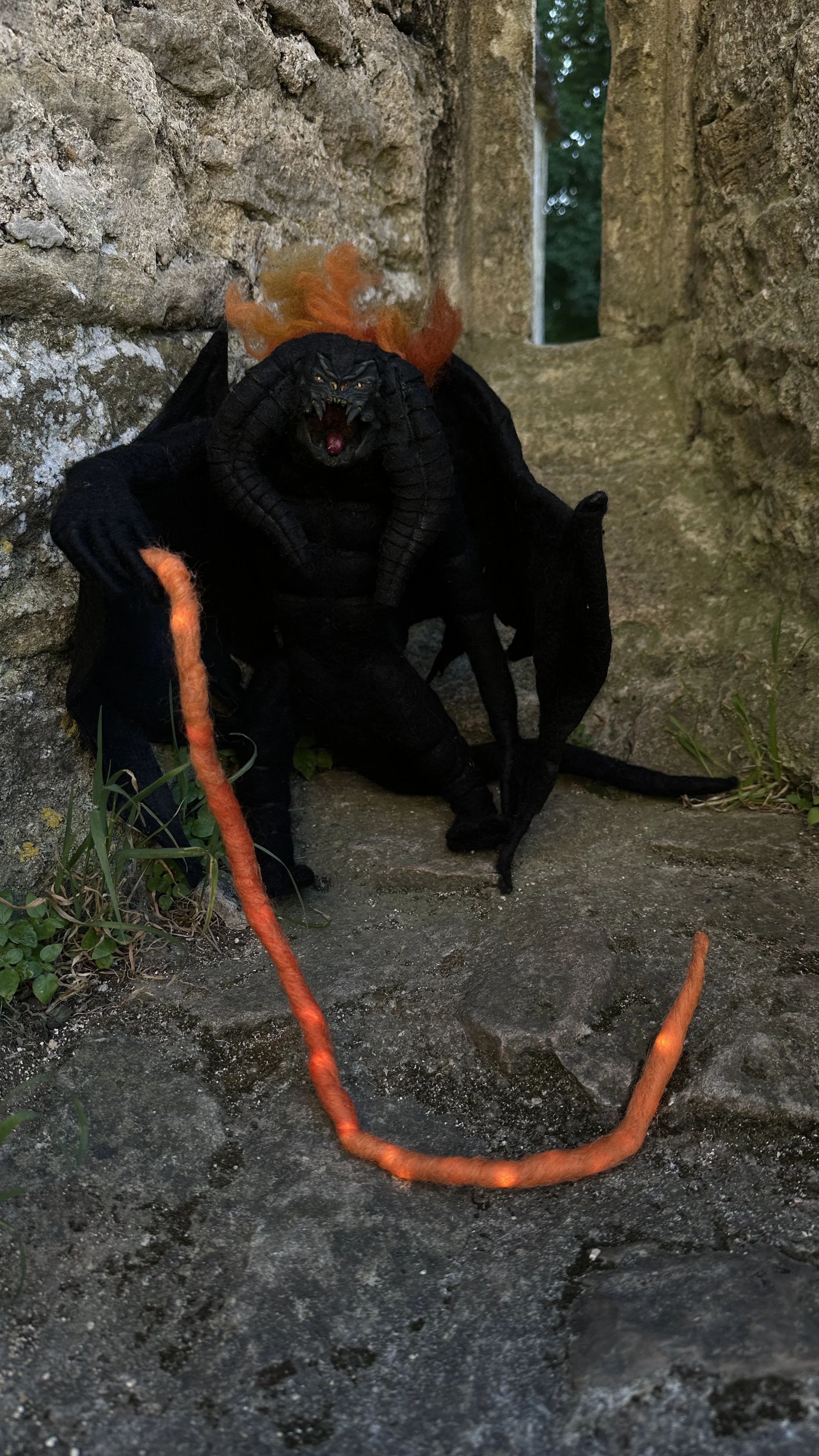 A detailed dragon figurine with black skin, horns, wings, and fiery orange hair, positioned outdoors between stone structures.