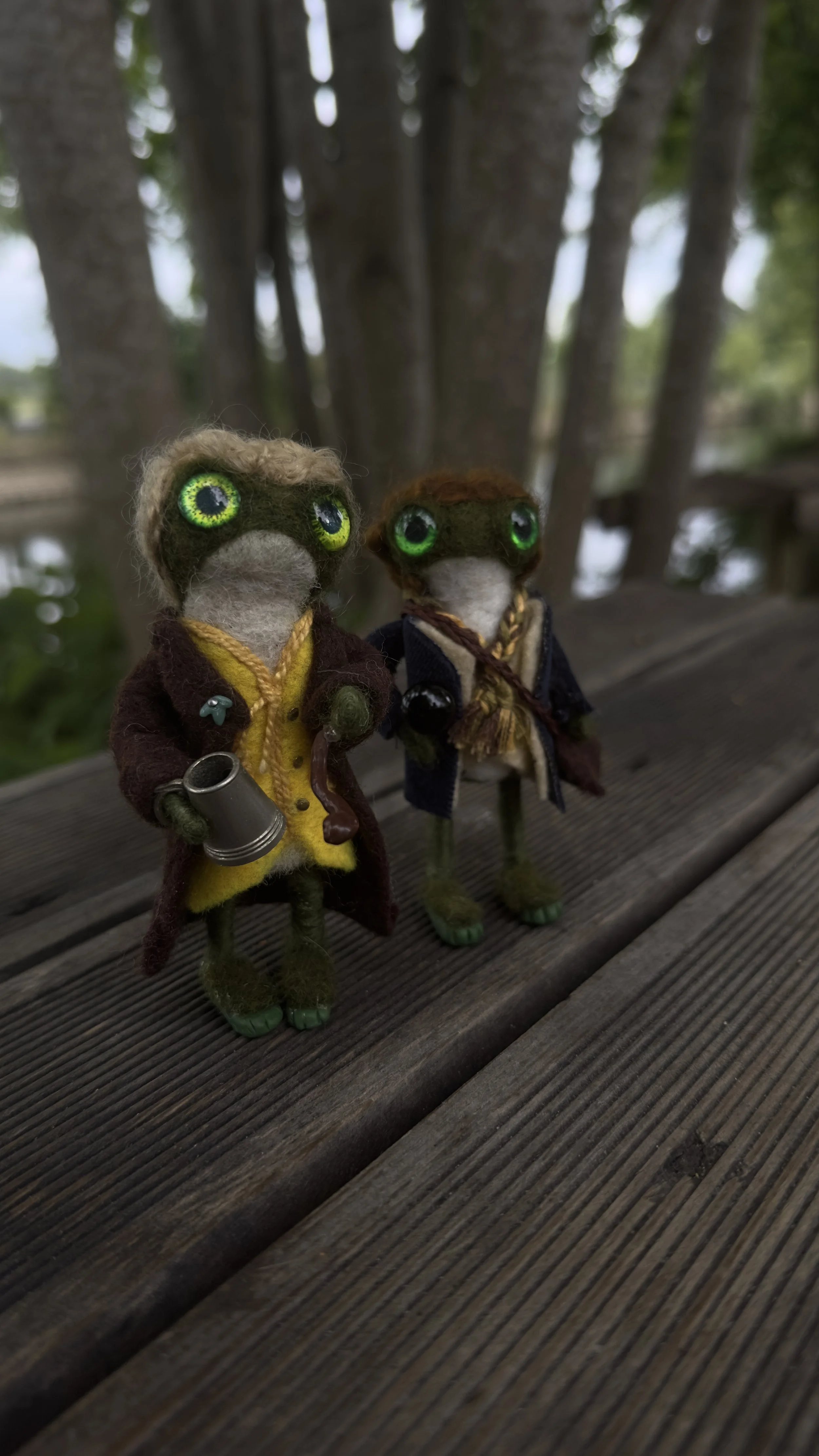 Two small handcrafted frog figurines with big green eyes dressed in detailed costumes, standing on a wooden surface outdoors with trees and water in the background.