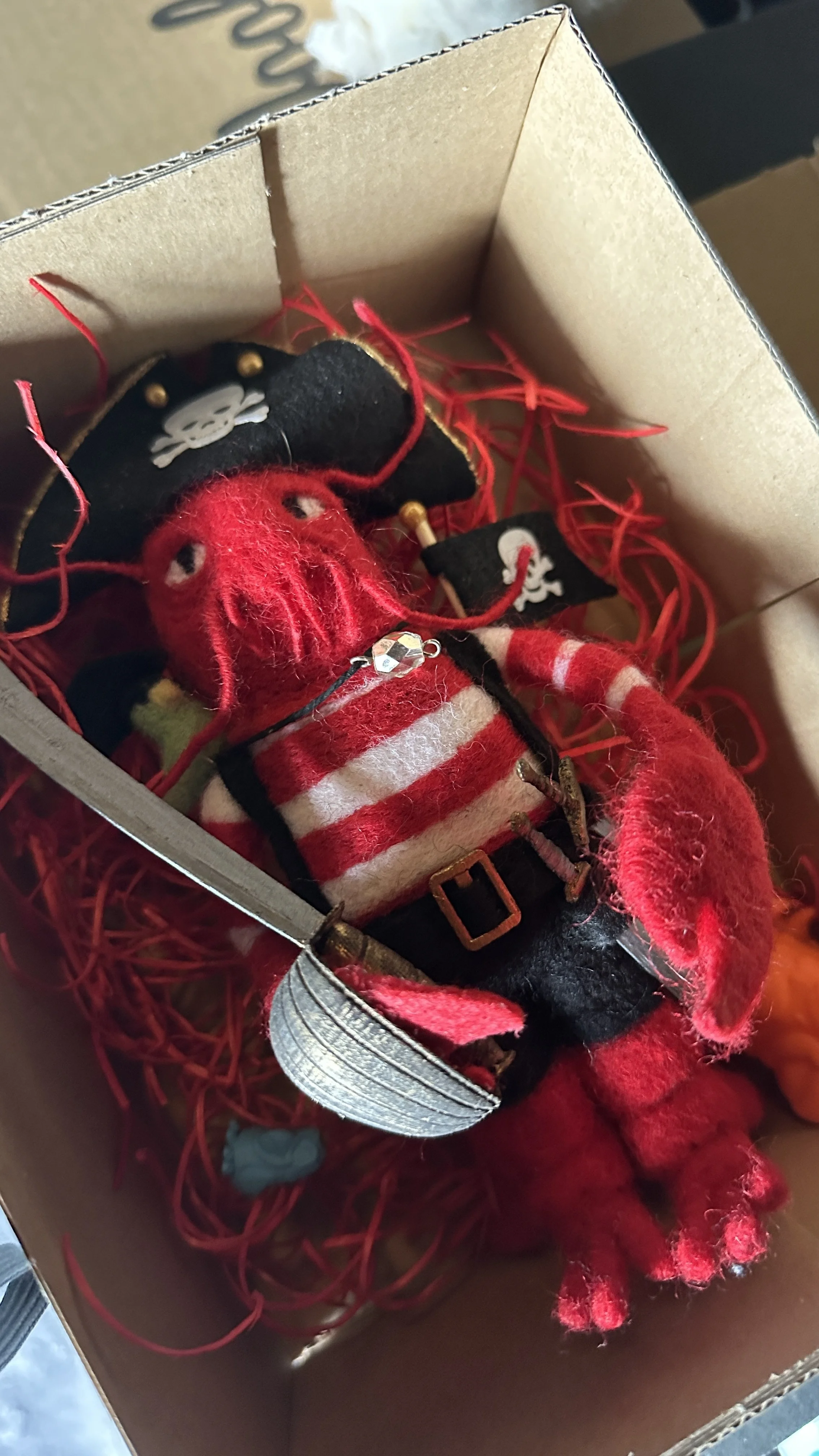 A felted doll lobster dressed as a pirate inside a cardboard box with red shredded paper. The lobster has red felt claws and legs, and wears a black eye patch with a skull and crossbones. It is also wearing a red and white striped shirt.