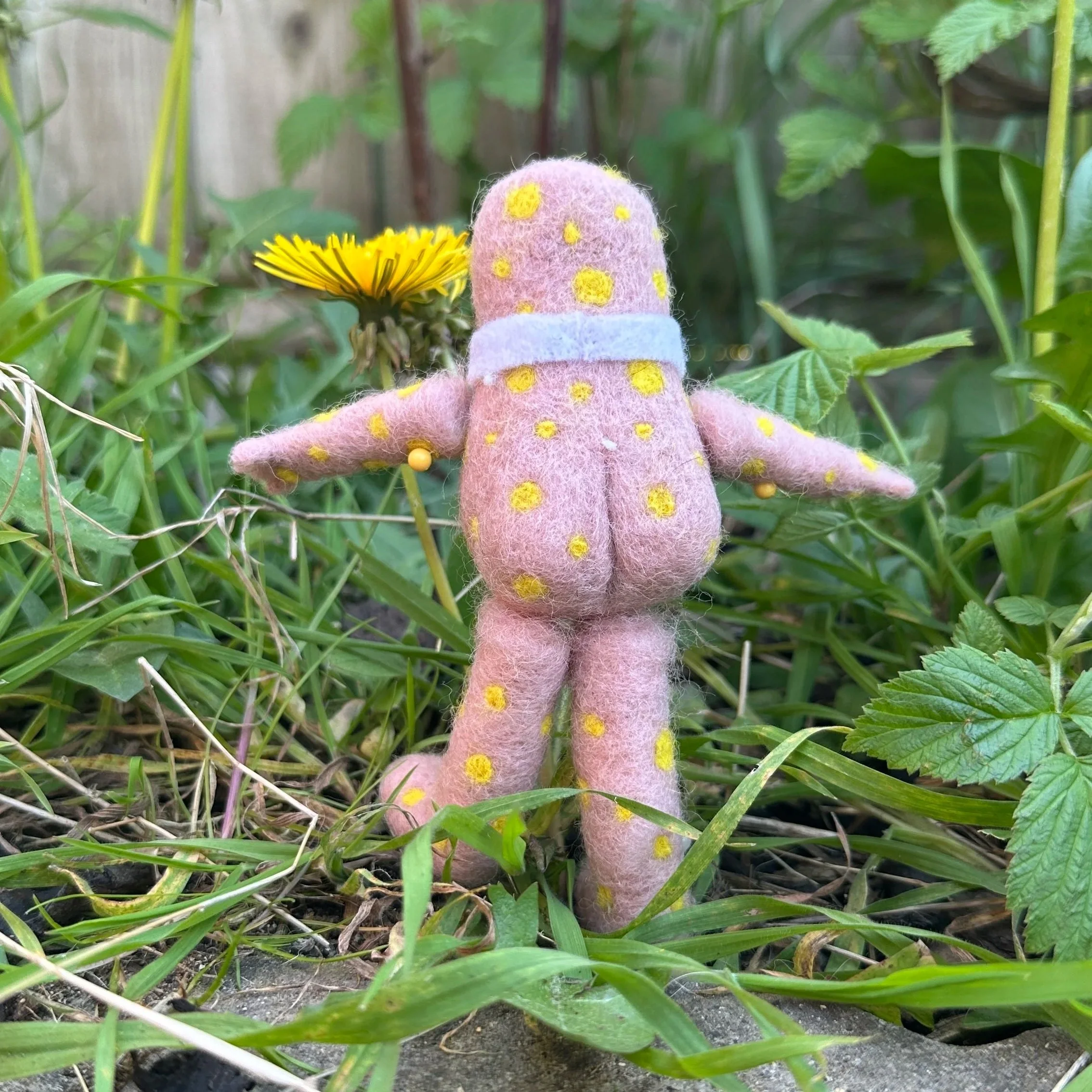 A felted doll with a pink body covered in yellow spots stands in a garden among green grass and plants, with a yellow dandelion flower in the background.